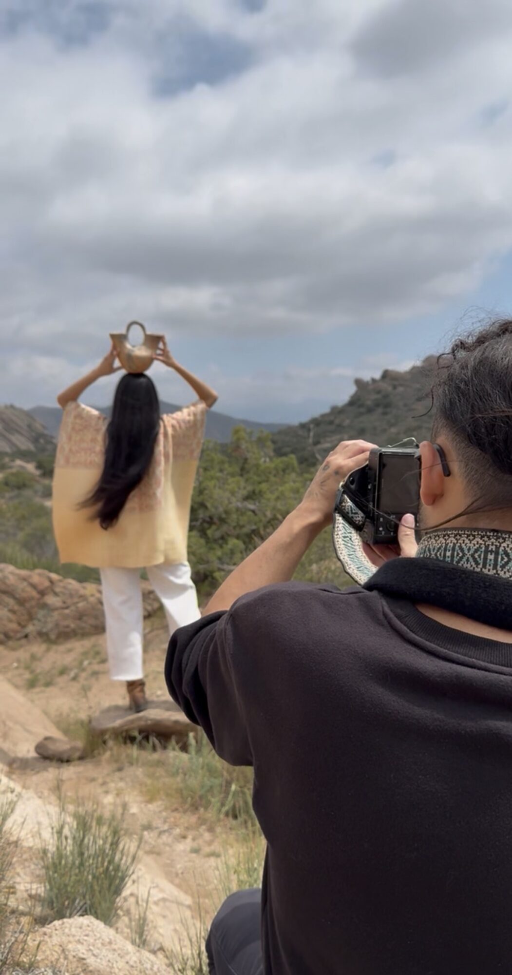 Person taking a photo of another person holding a hat outdoors with mountains and cloudy sky in background.