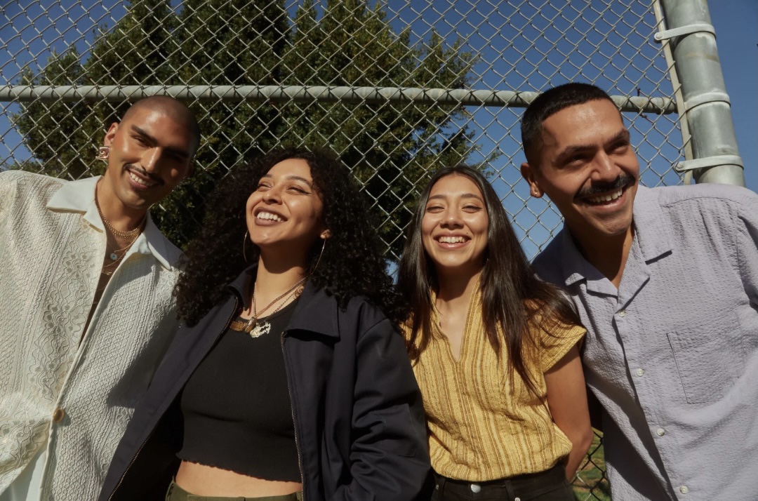Four people smiling outdoors in front of a chain-link fence, sunny day, clear blue sky.