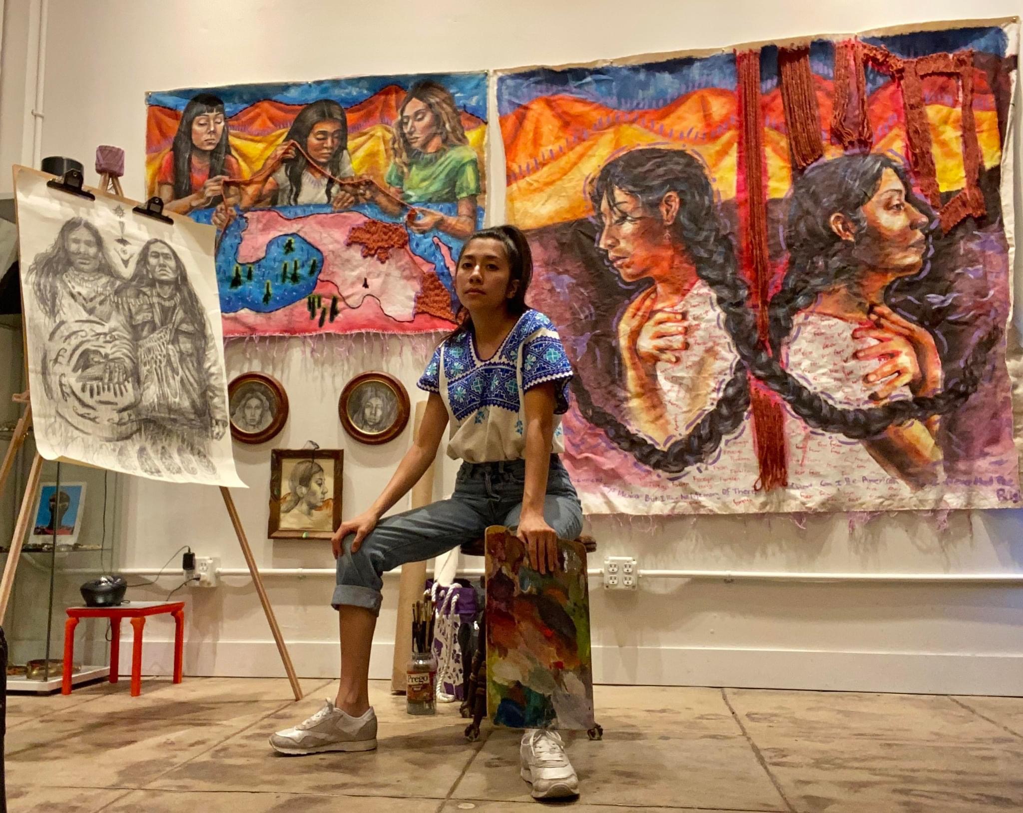 Young woman sitting in front of colorful paintings and a sketch on an easel, holding a paintbrush and palette.