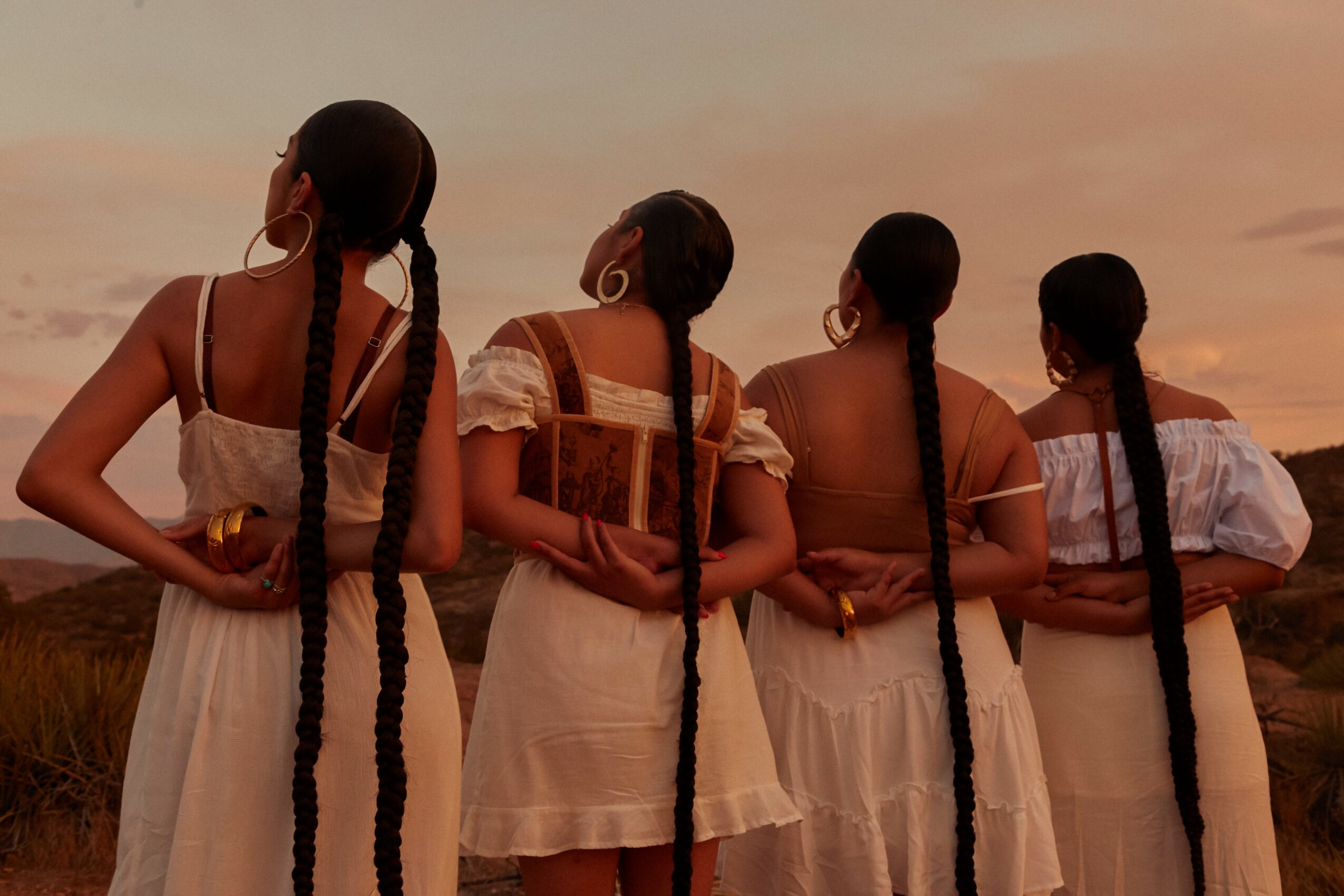 Four women with long braided hair stand outdoors at sunset, facing away, wearing white dresses, holding a book and a box.