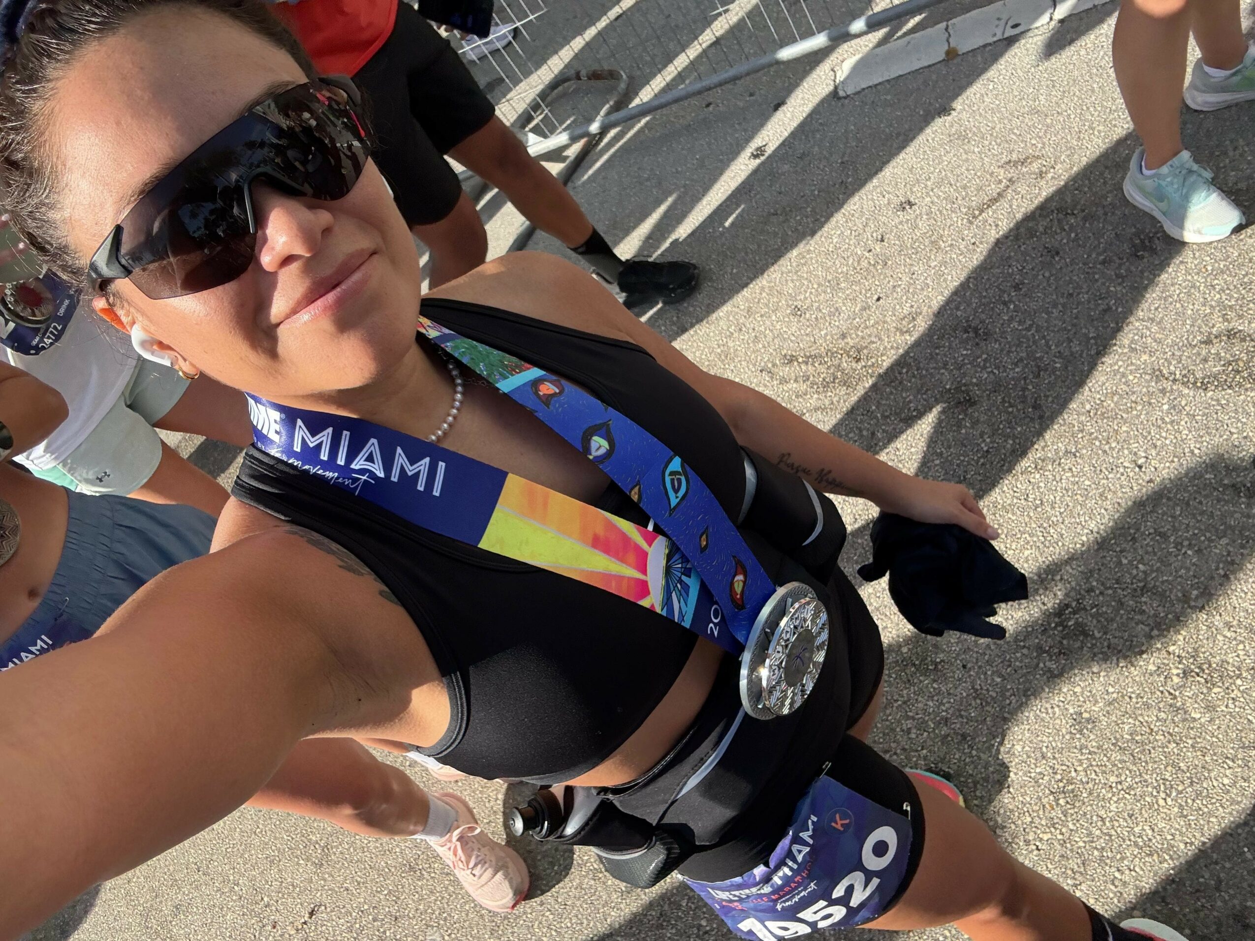 Woman wearing sunglasses, a medal, and a race bib, taking a selfie outdoors with shadows on the ground.