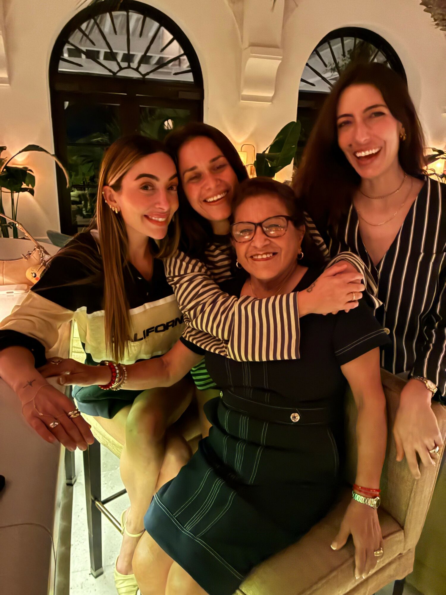 Four women sitting together, smiling, in a cozy indoor setting with arched windows and plants.