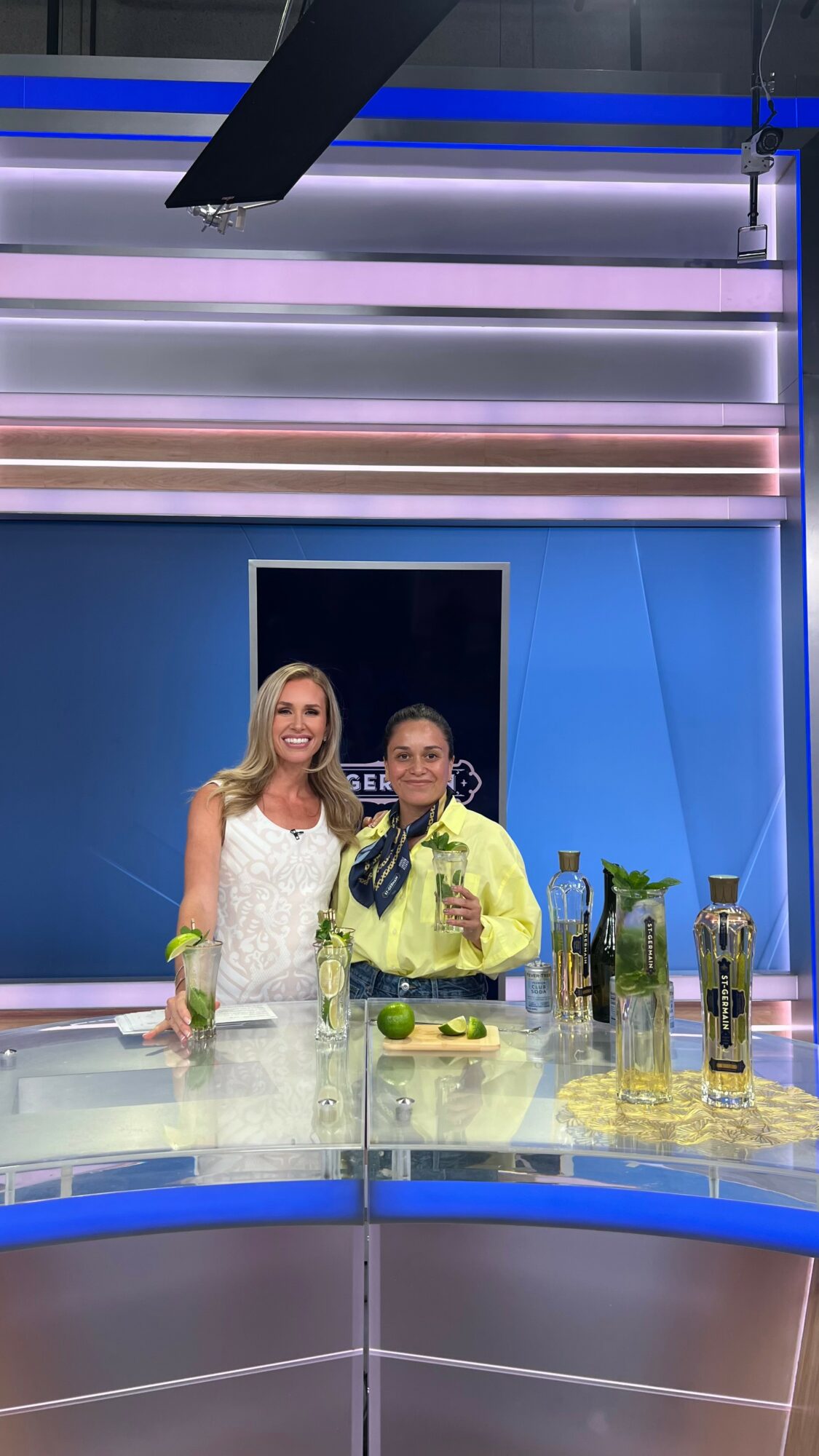 Two women stand behind a bar with bottles and limes, smiling at the camera, in a brightly lit studio.