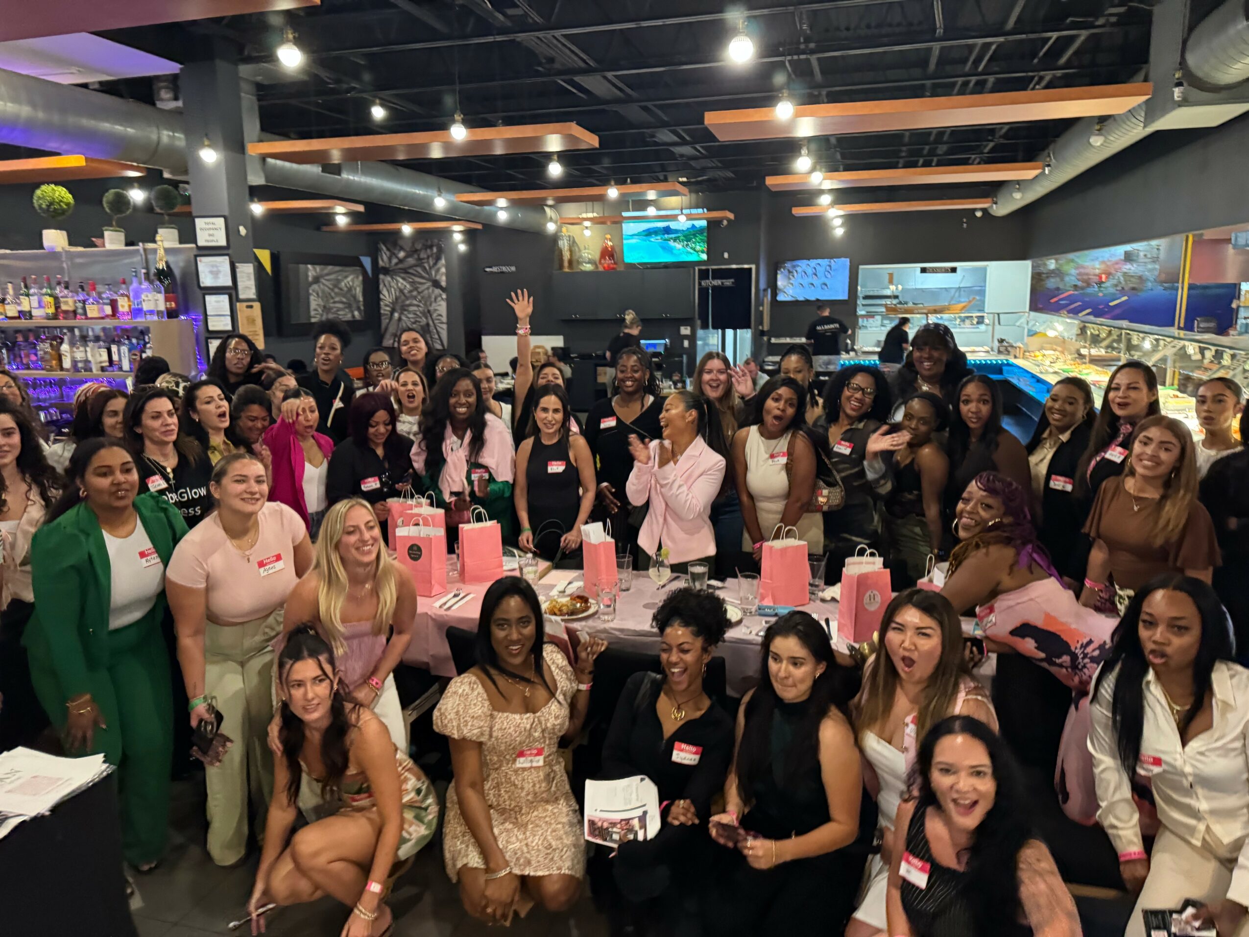 Group of women gathered in a bar or restaurant, some holding pink gift bags, smiling and celebrating together.