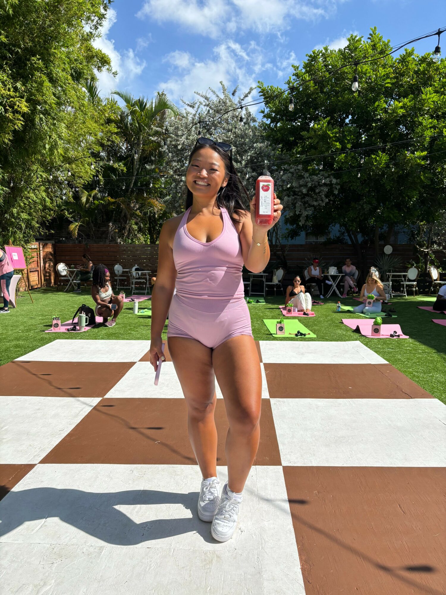 Young woman in pink athletic outfit standing on checkered outdoor surface, smiling, with trees and people in background.