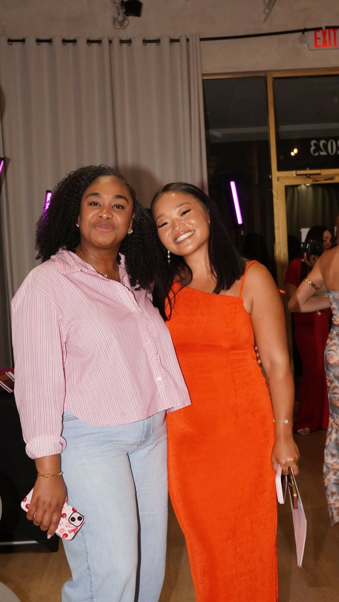 Two women standing indoors, one in a pink shirt and light blue pants, the other in an orange dress, smiling.