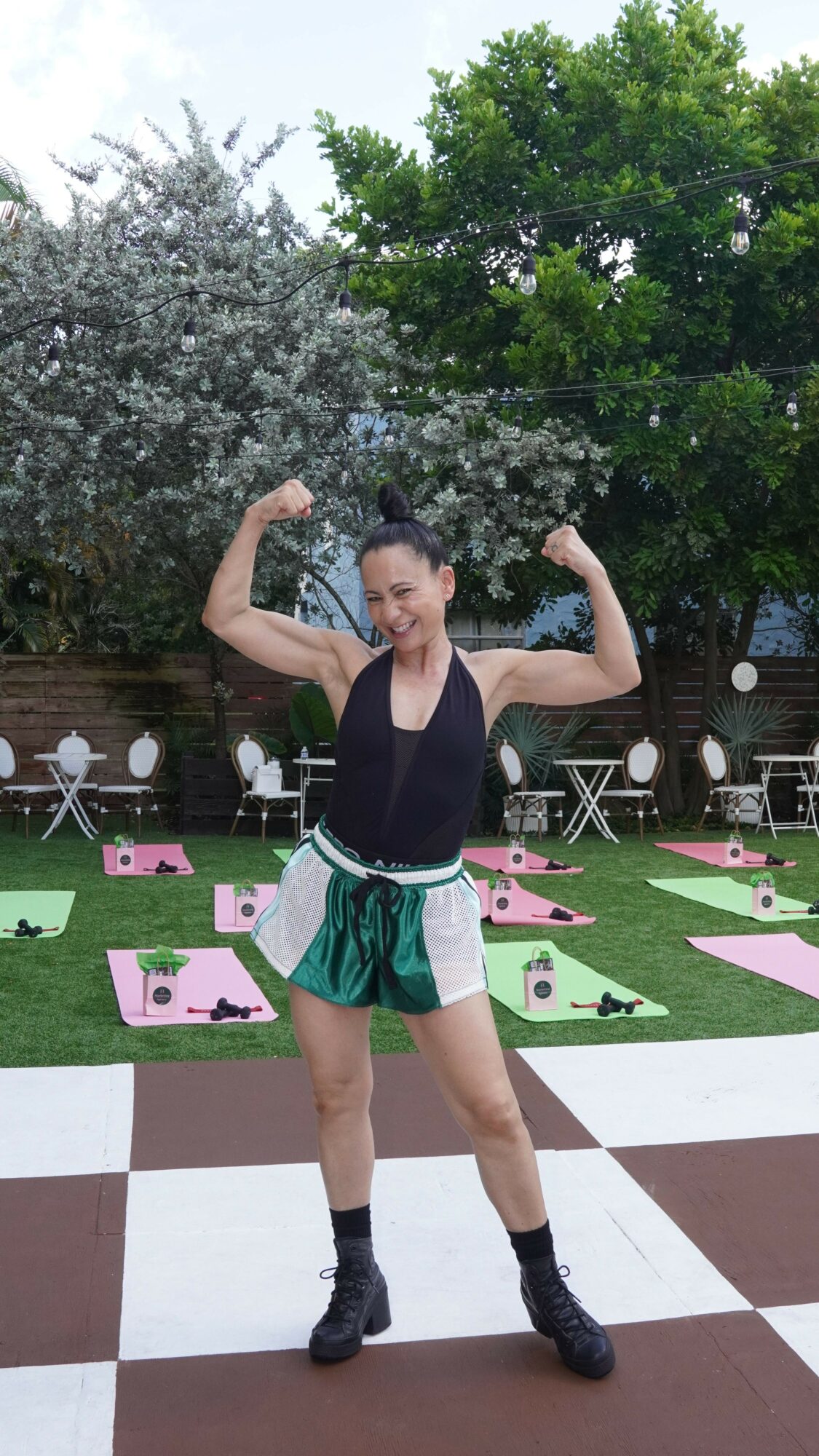 Person standing outdoors on checkered floor, flexing arms, surrounded by trees and pink and green decorations.