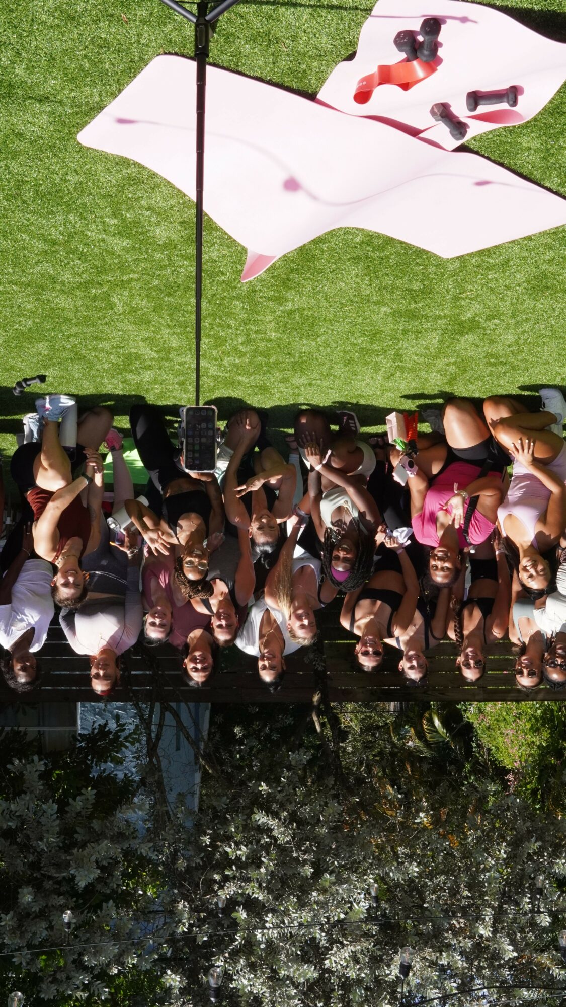 Group of people gathered outdoors on grass, some sitting and some standing, with trees and a building in background.