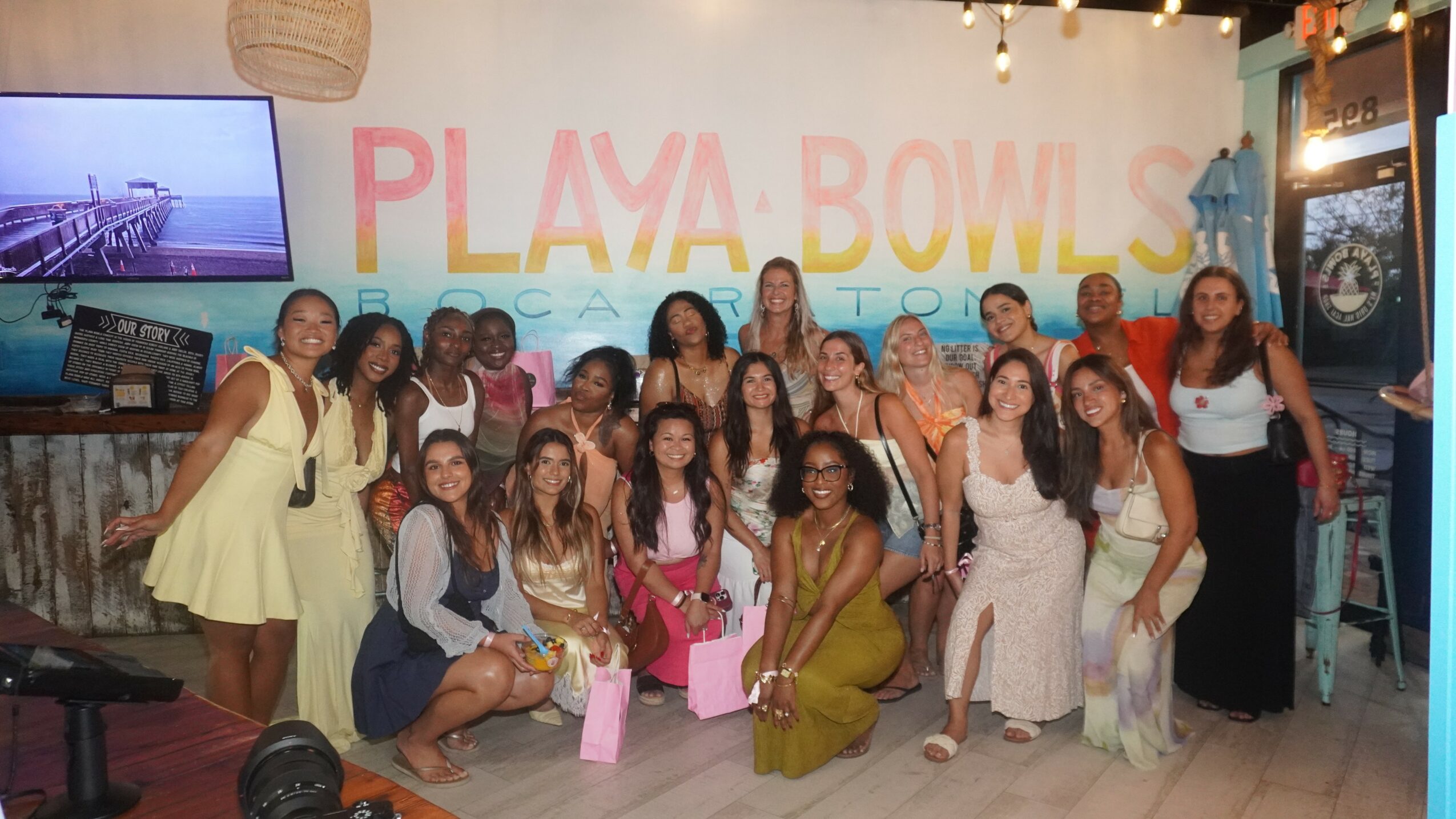 Group of women posing in front of a wall with colorful text, some holding pink bags, indoors.