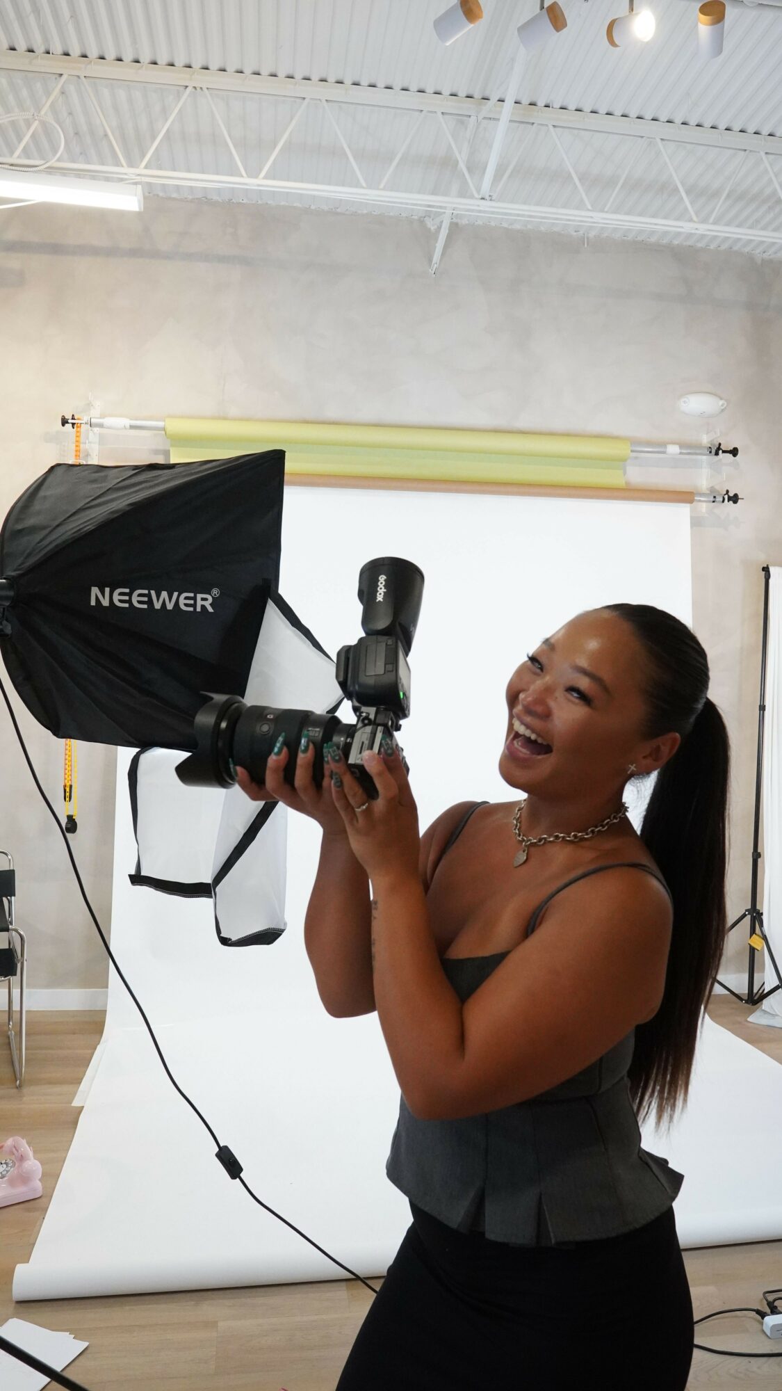 Woman holding a camera in a photography studio, smiling, with lighting equipment and a backdrop in the background.