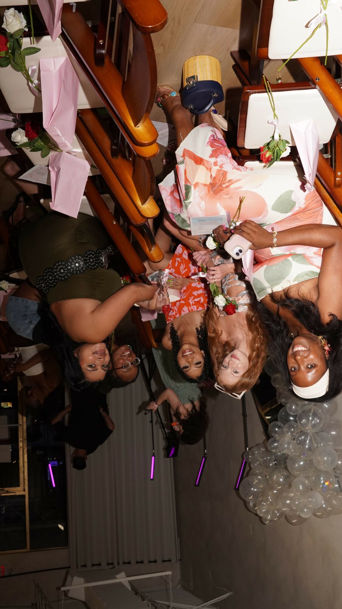 Group of women sitting at a table with flowers, drinks, and pink boxes, in a dimly lit room.