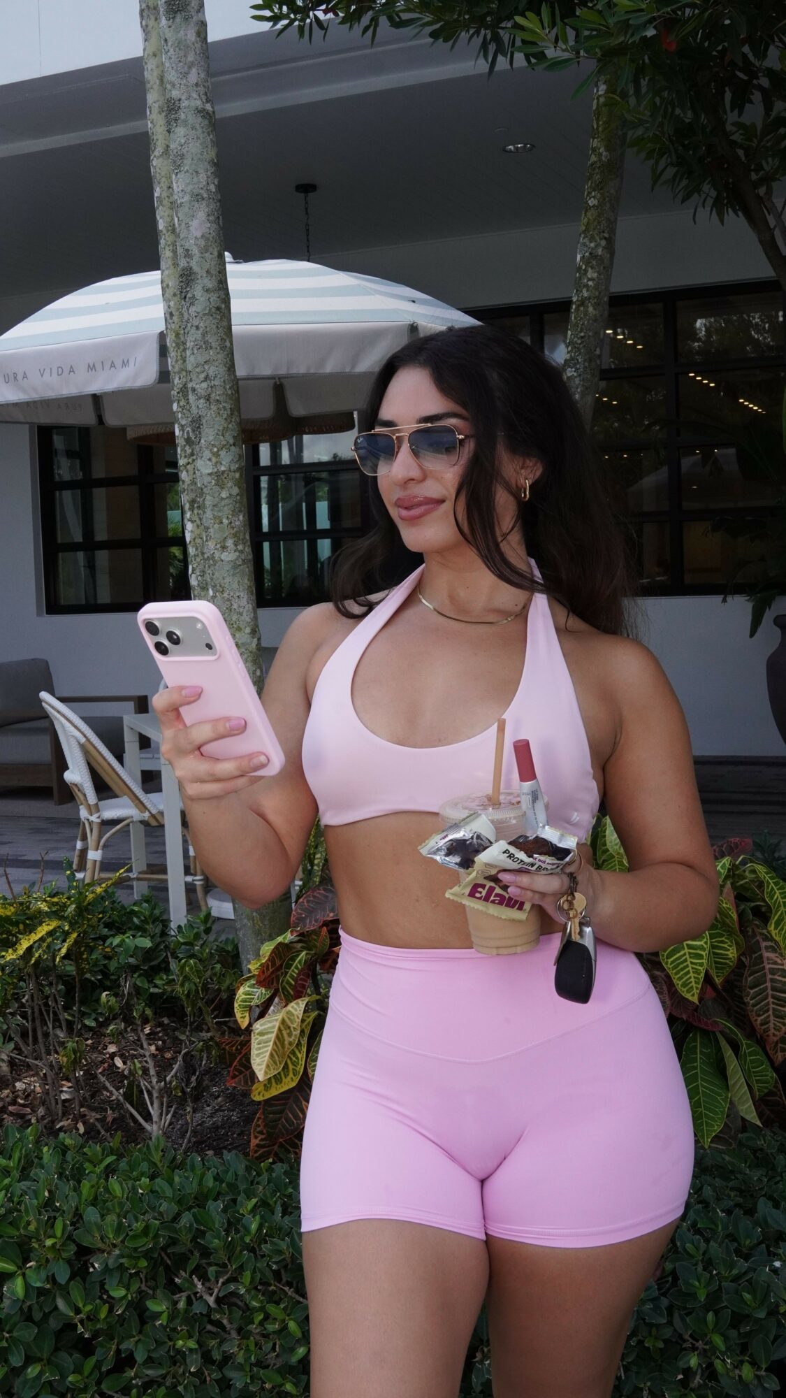 Woman in pink outfit holding a phone, sitting outdoors near a tree, with a building and car in background.