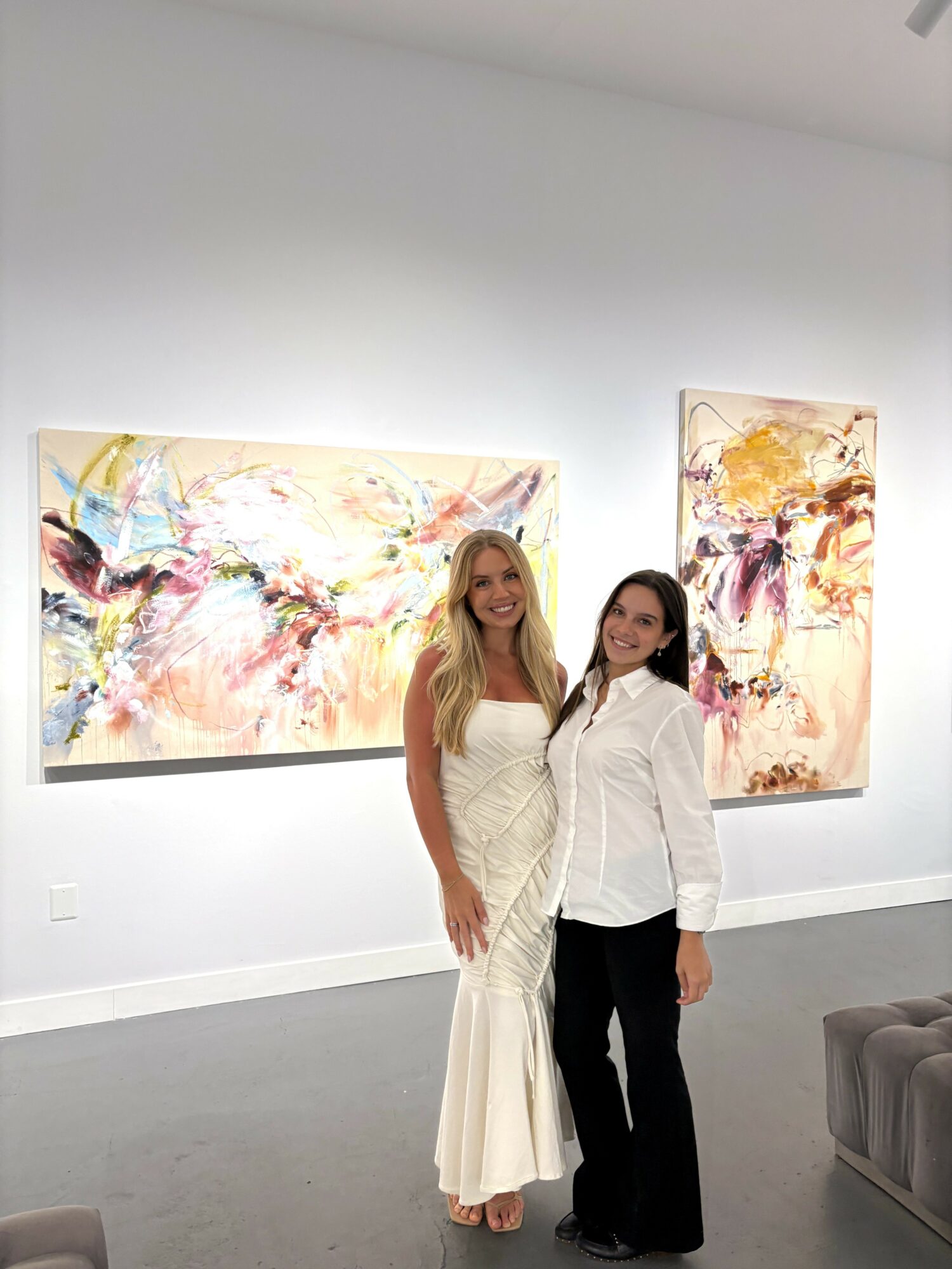 Two women stand in front of colorful abstract paintings in an art gallery, smiling at the camera.