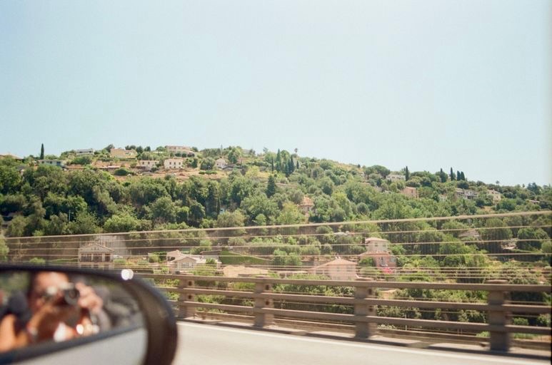 Landscape view of a hillside with buildings and trees, seen from a moving vehicle with a side mirror reflecting part of the scene.