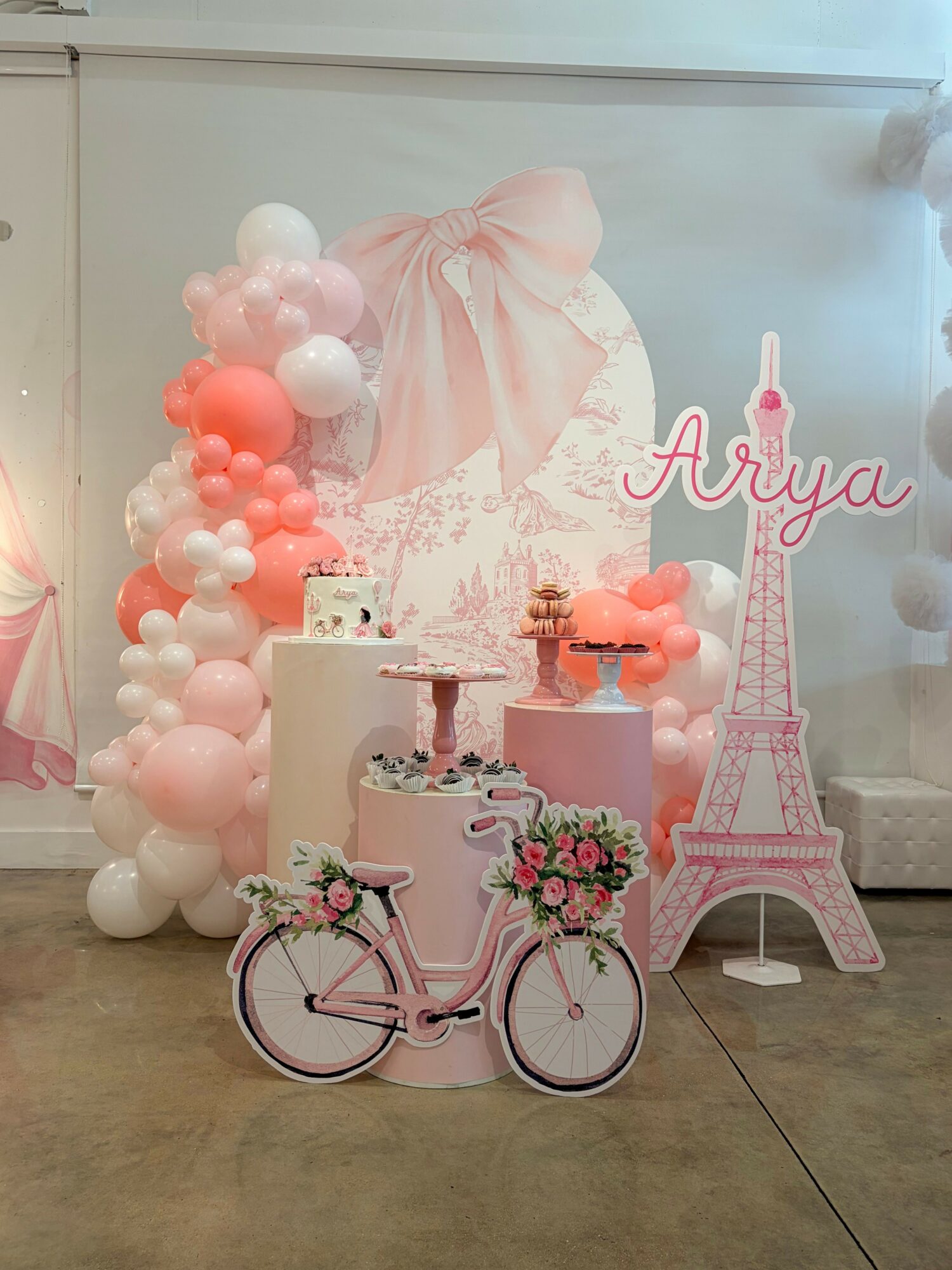 Decorative pink and white birthday setup with balloons, a cake, a bicycle, and a pink Eiffel Tower replica, celebrating a girl named Thuy. Alt text word count: 37
