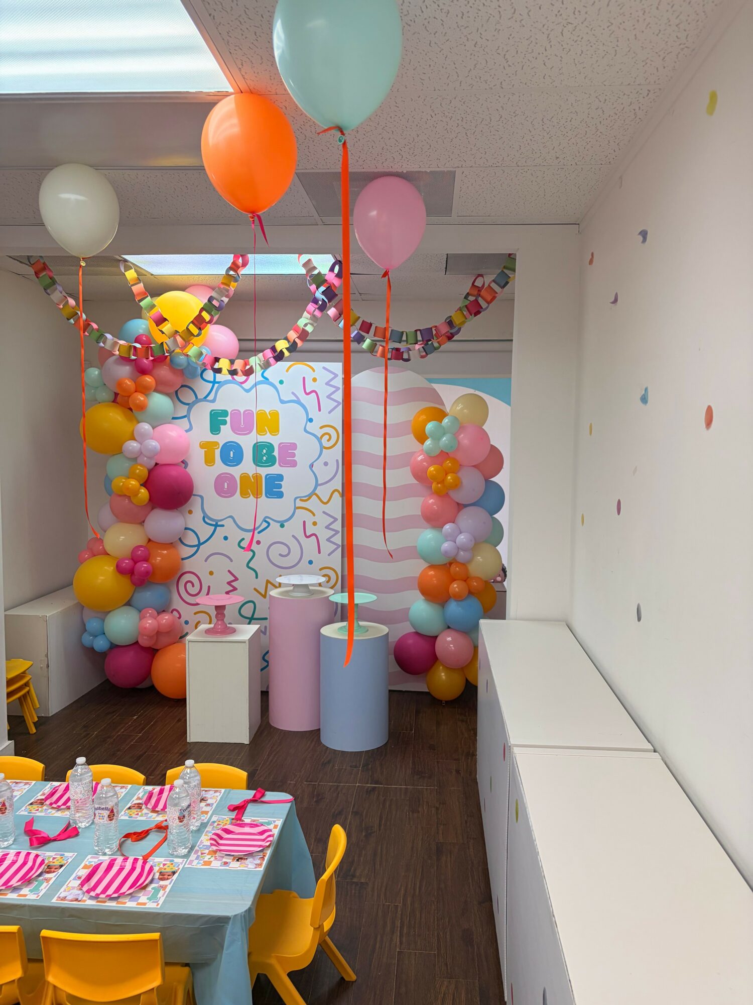 Party room decorated with balloons, streamers, and a colorful backdrop, featuring a table with cupcakes and party supplies.