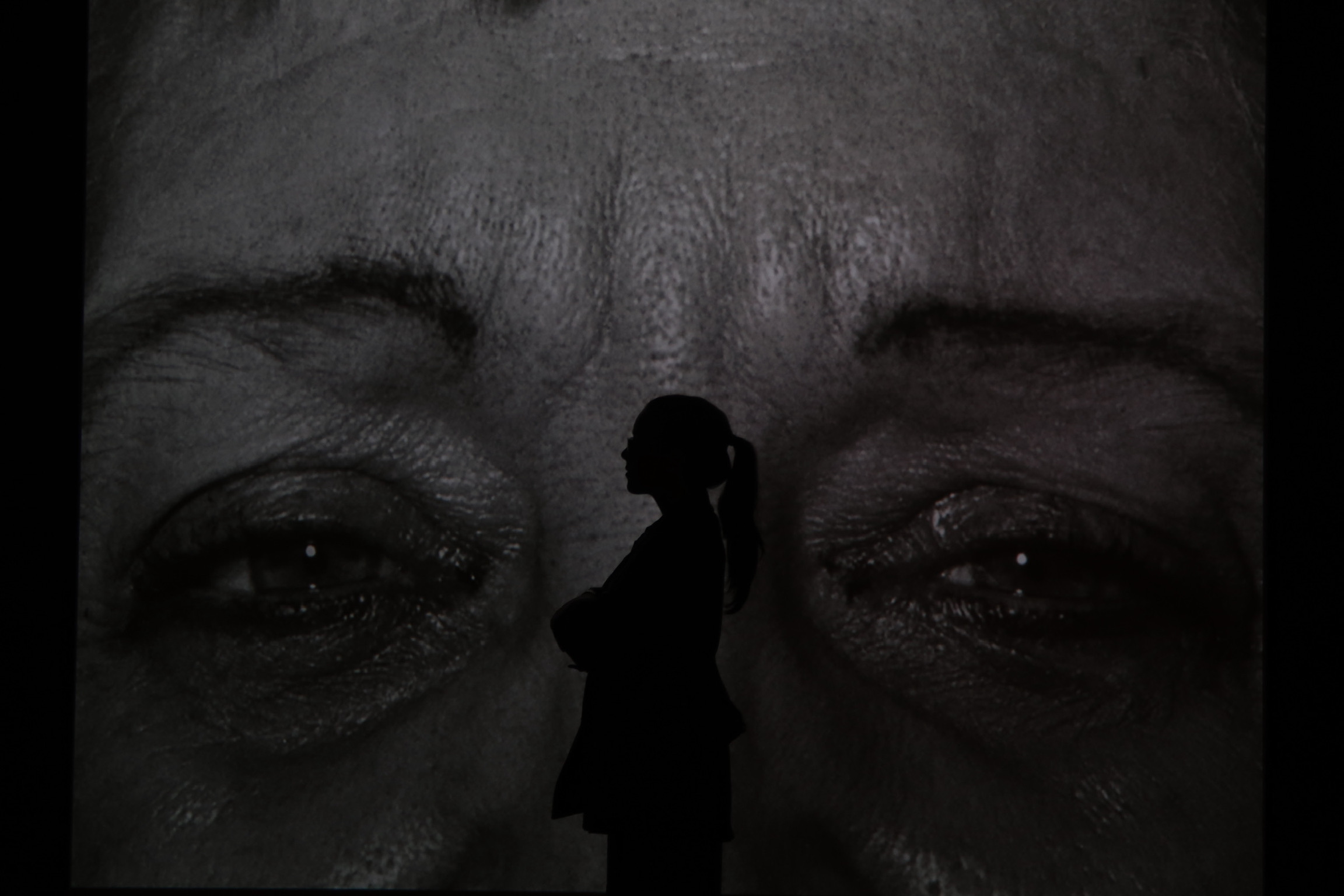 Silhouette of a person standing in front of a large, close-up image of a face with closed eyes, in black and white.