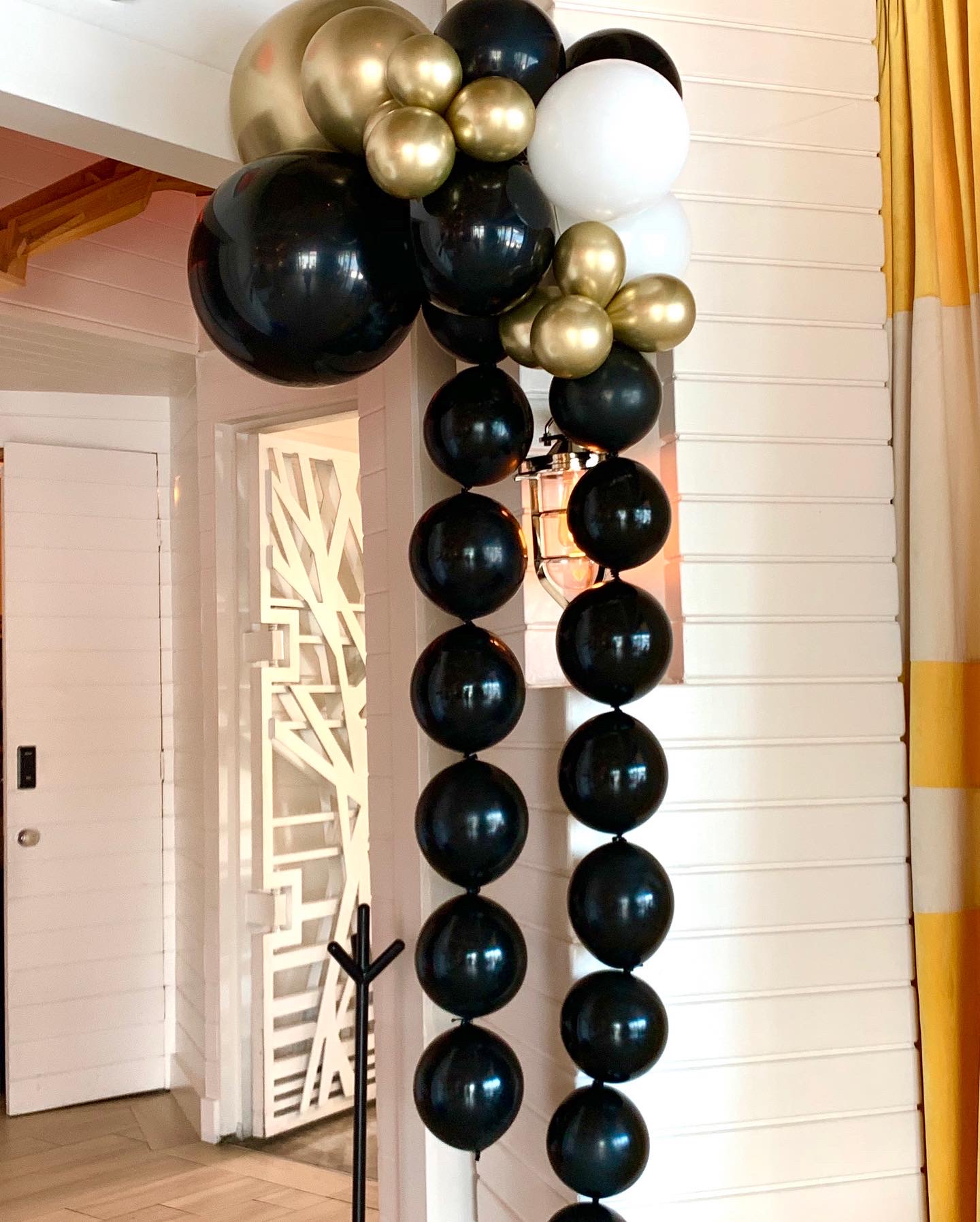 Balloon arch with black, gold, and white balloons arranged vertically indoors, near a white wall and door, with yellow curtains nearby.