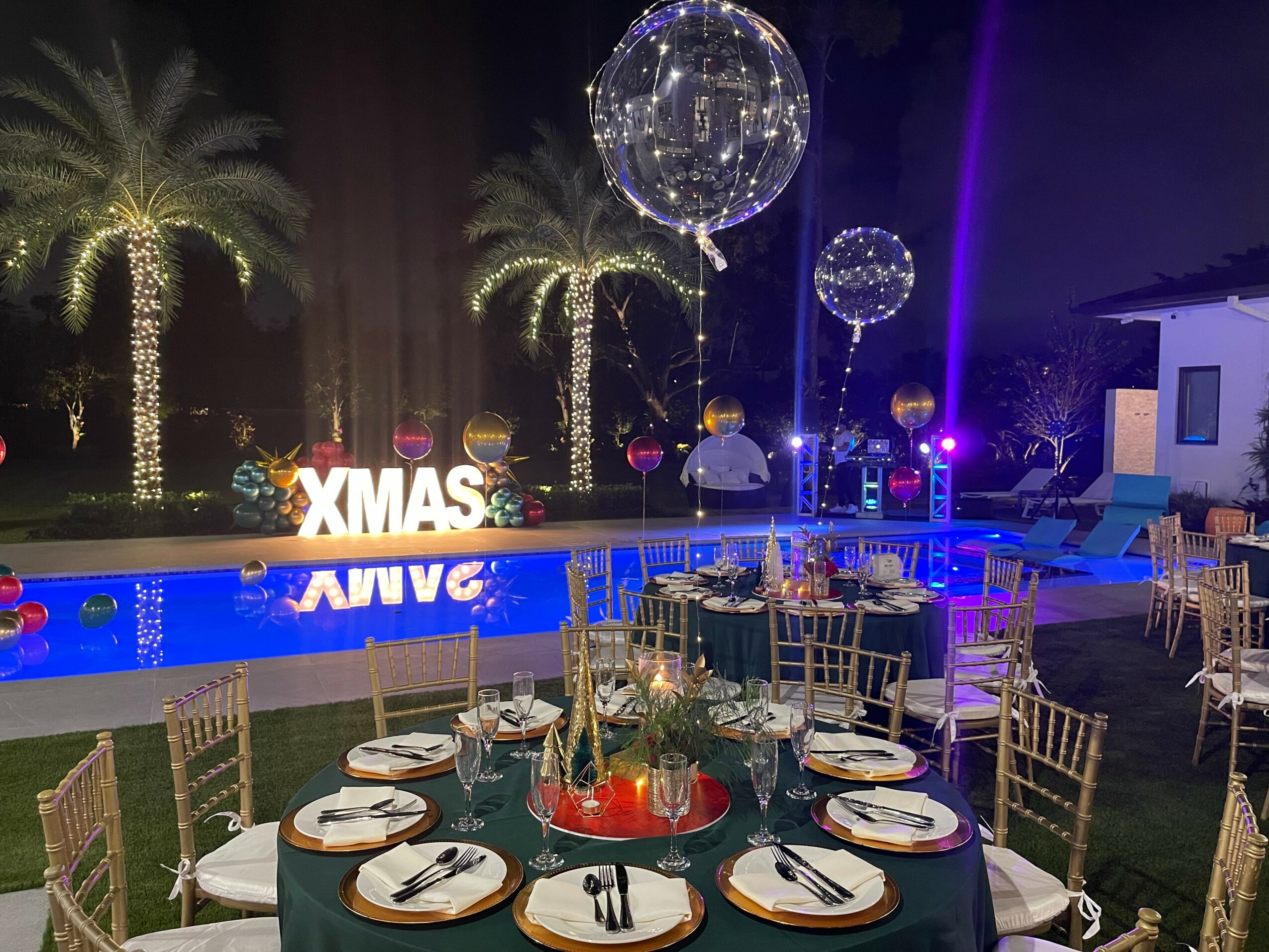 Party setup with tables, chairs, and decorations near a pool, illuminated by colorful lights and large hanging balloons.