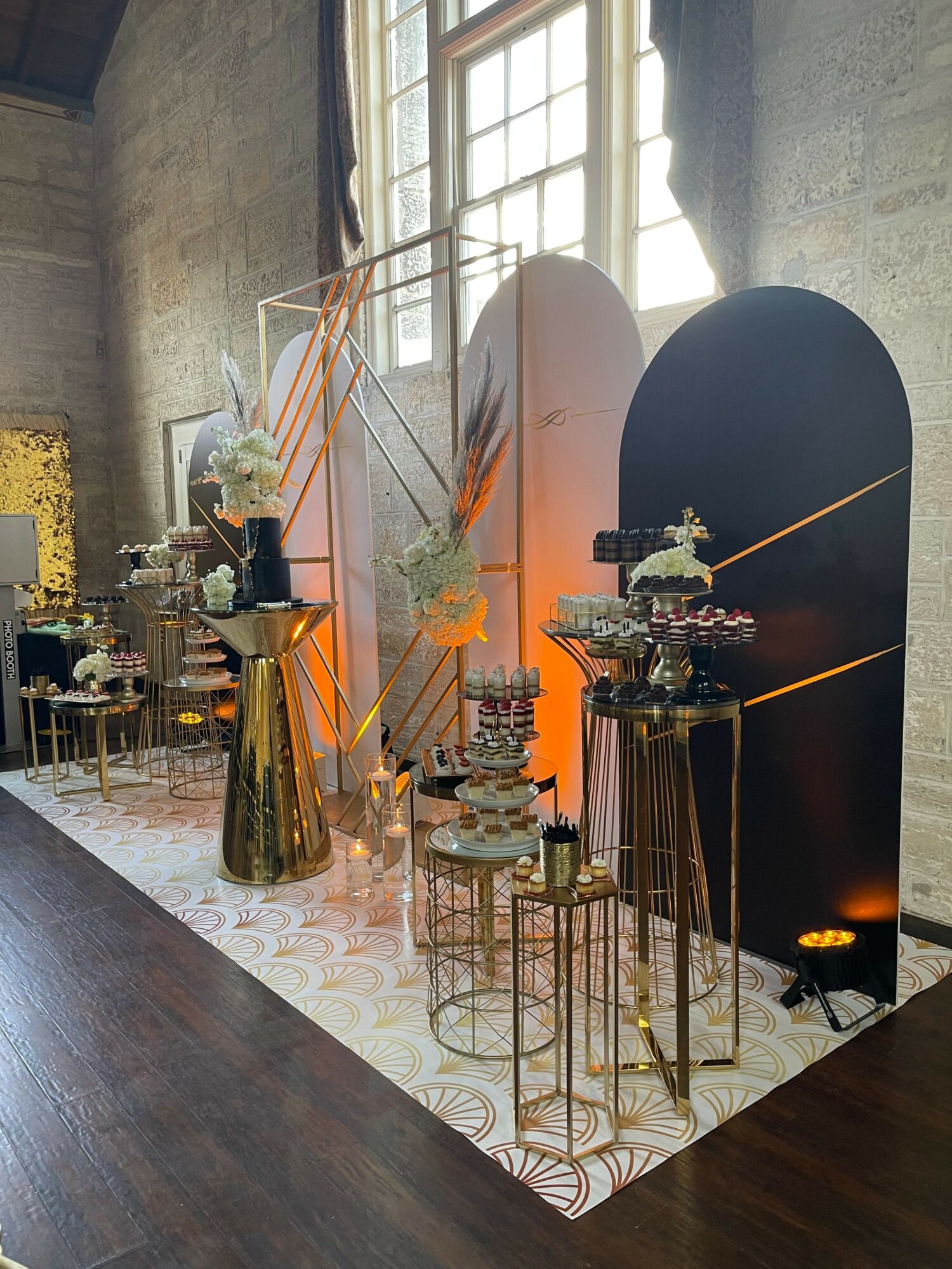 Decorated table with cakes, flowers, and gold accents, set against a window and wall, with a black backdrop and orange lighting.