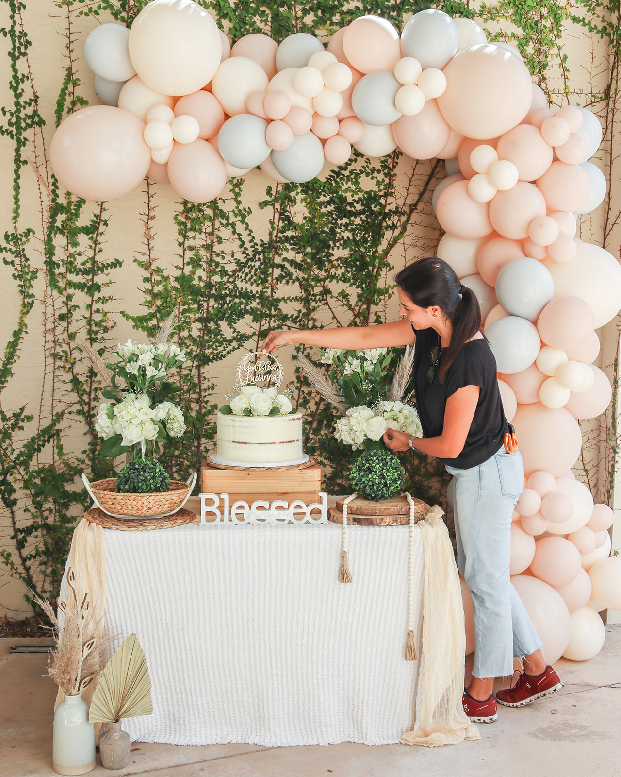 Woman in black shirt and jeans reaching for a cake on a decorated table with balloon arch backdrop.