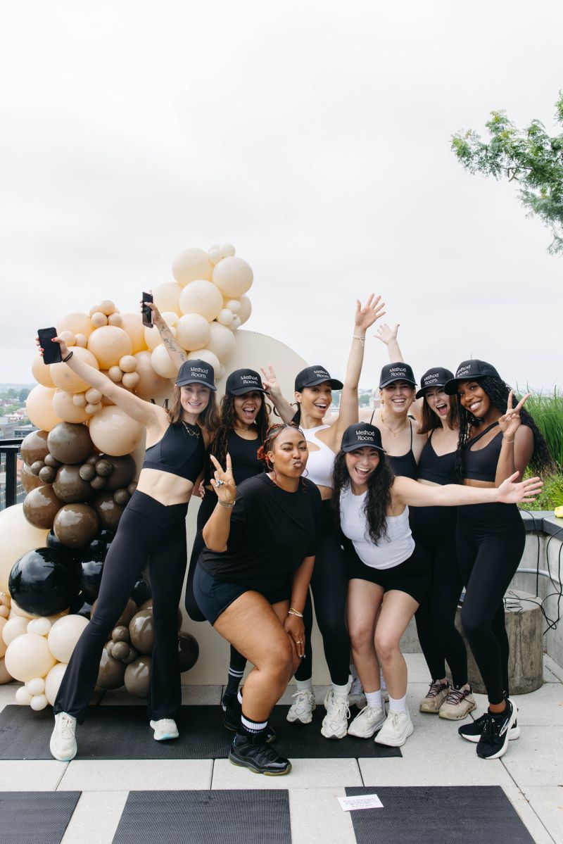 Group of seven women posing outdoors with balloon decorations, some making peace signs, smiling, and waving.