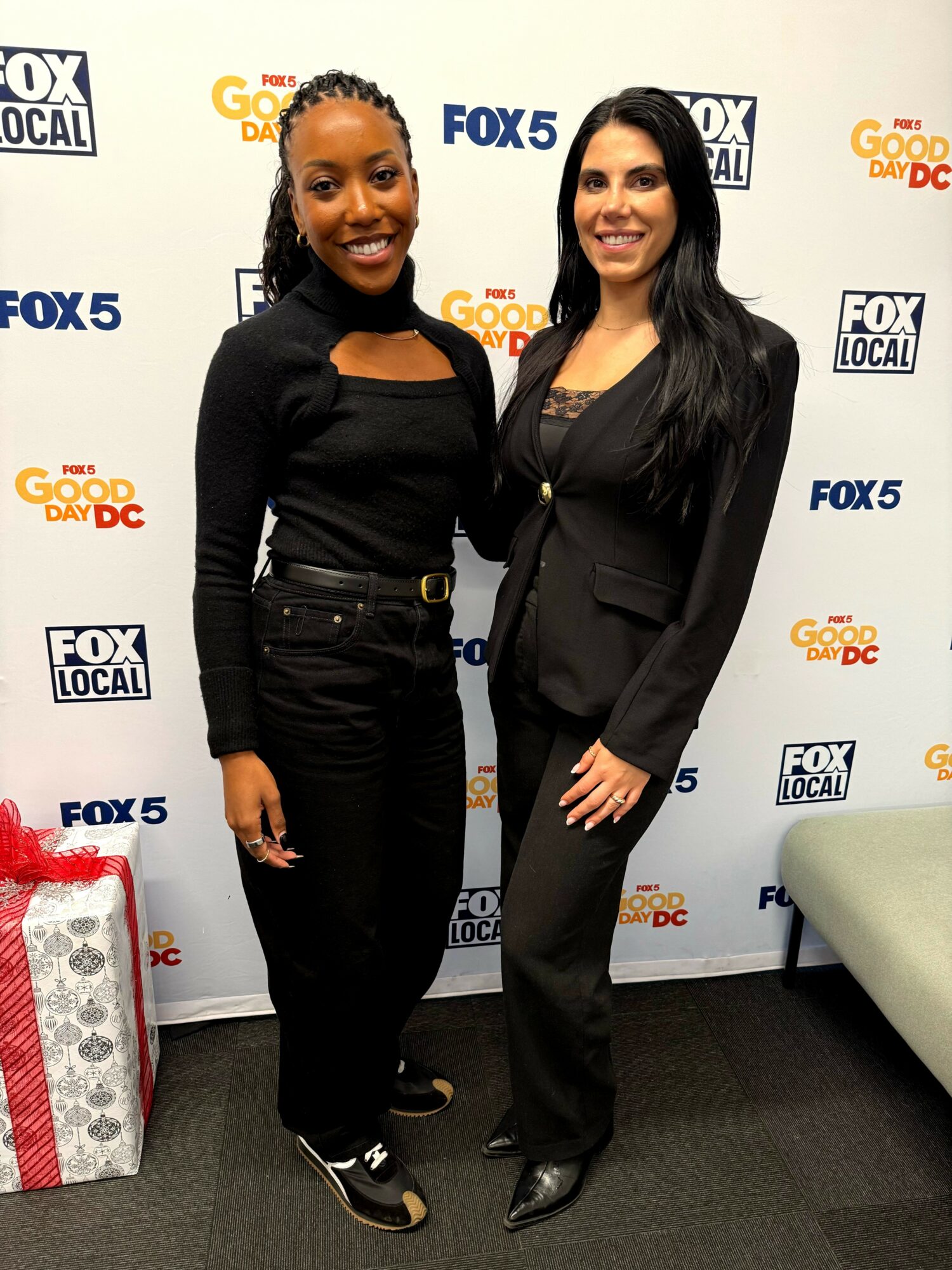 Two women standing together in front of a backdrop with TV station logos, smiling, one in black and one in dark suit.