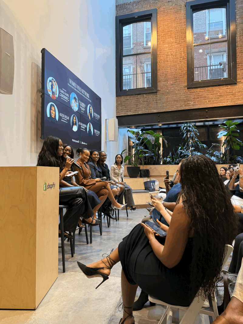 People seated in a modern indoor space with large windows, a screen on the wall, and plants, some using phones.