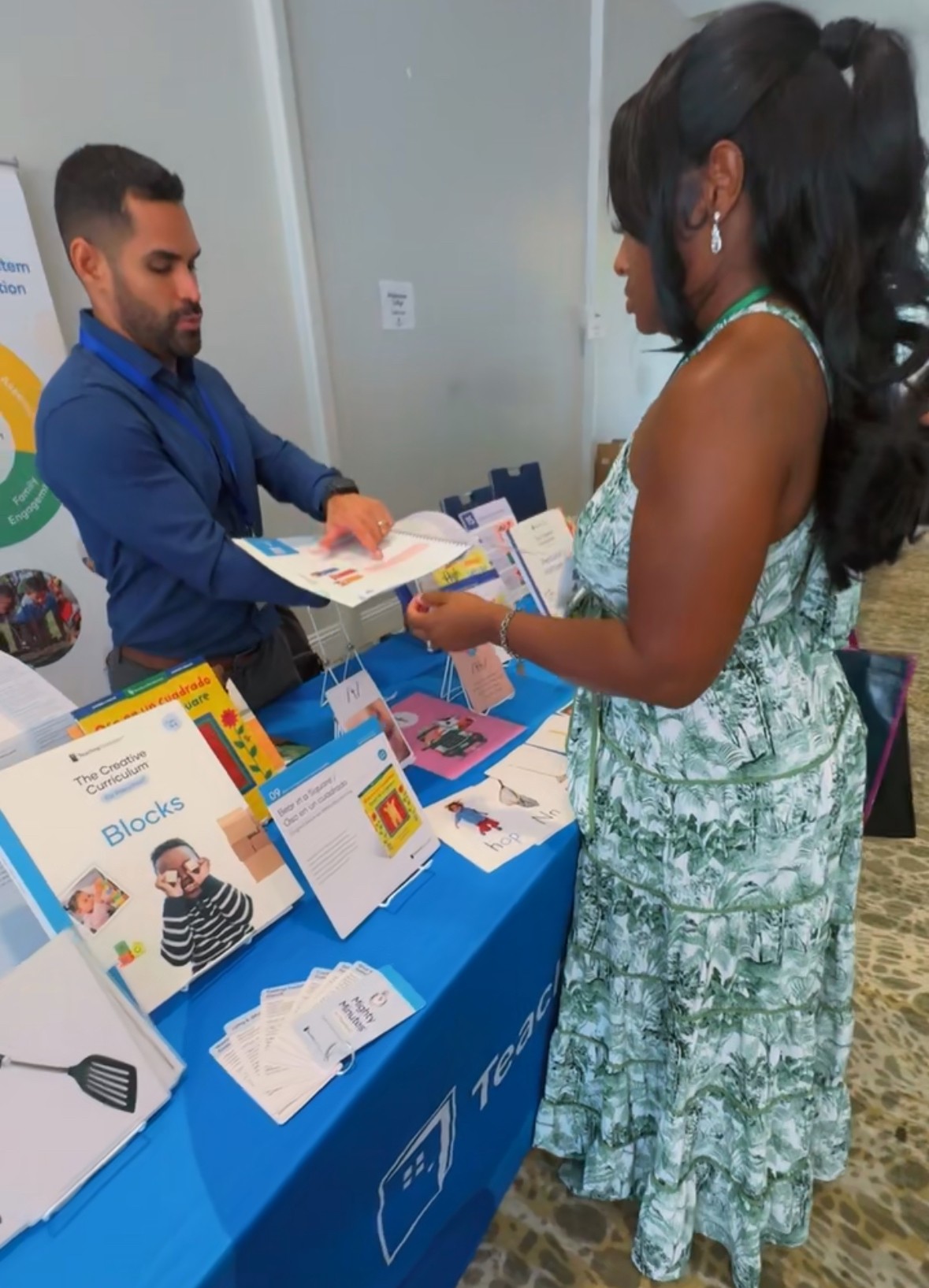 A woman in a floral dress interacts with a man at a booth displaying informational materials, in an indoor setting.