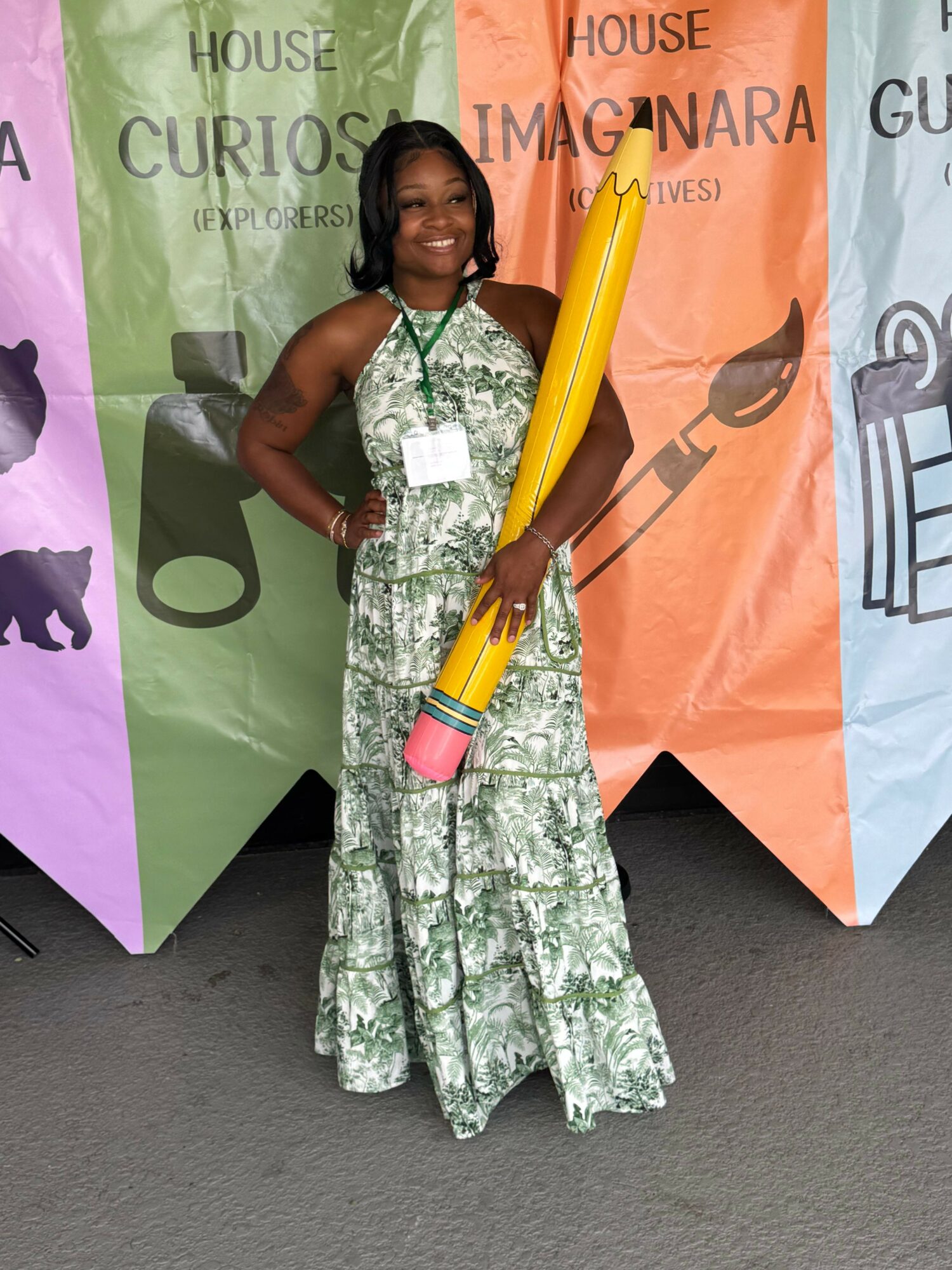 Child in a green and white patterned outfit holding a large yellow pencil prop, standing on a gray surface with colorful banners behind.