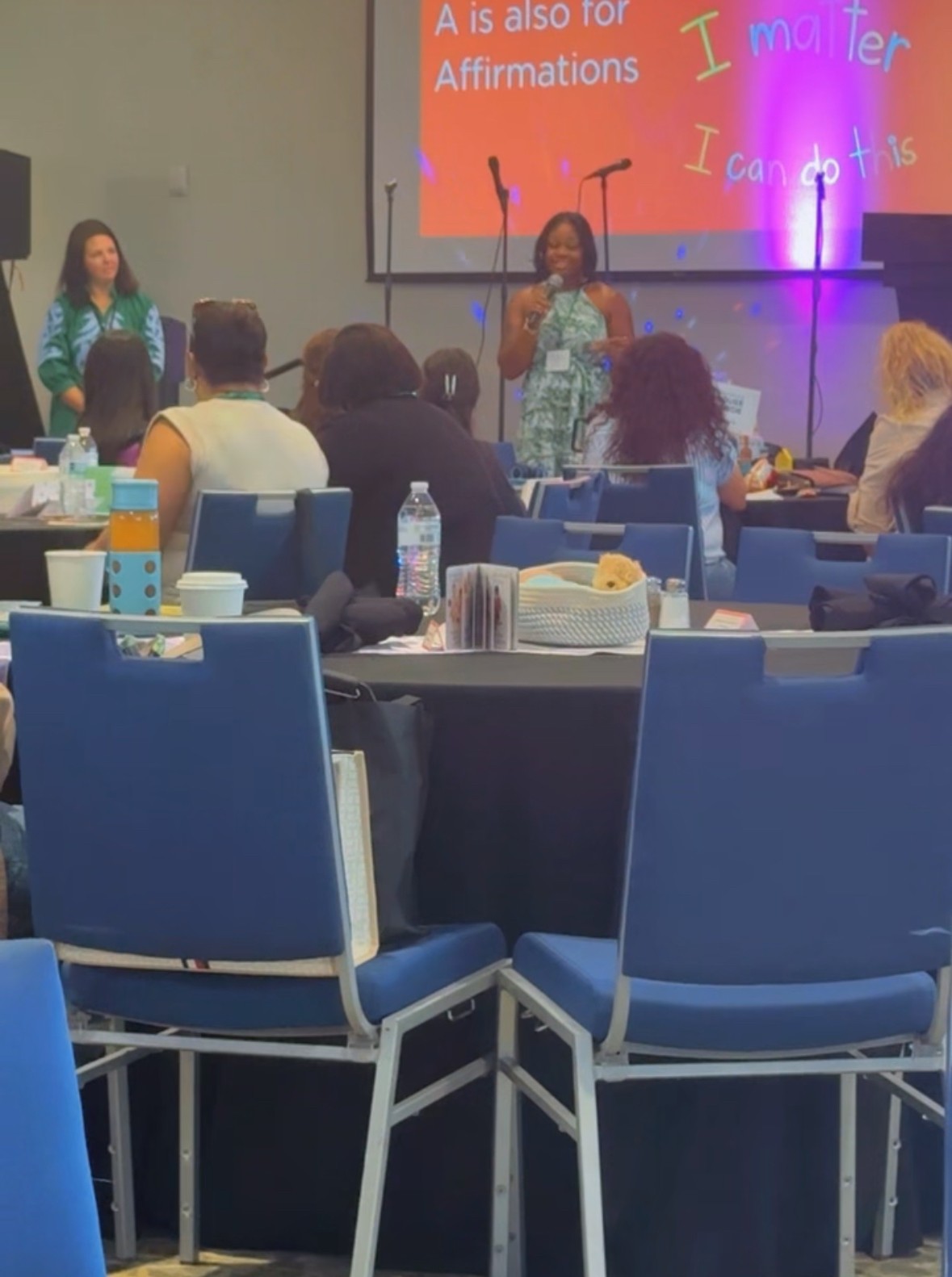 Two women speak at a conference with a large screen behind them displaying colorful text. Audience seated at tables in front.