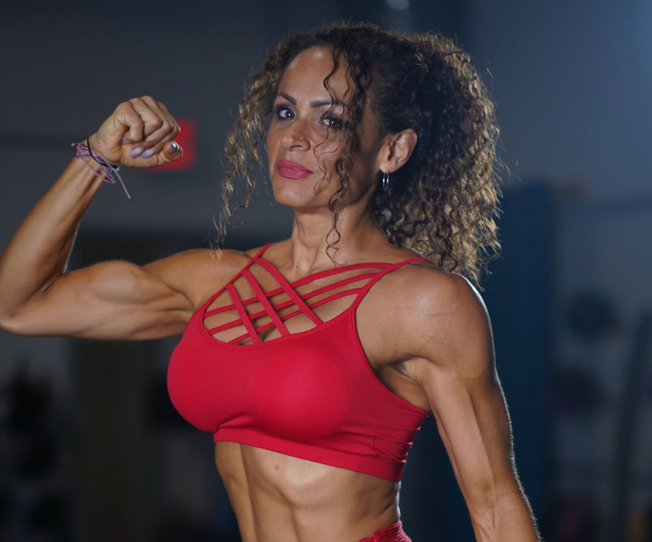 Woman flexing her arm muscles in a red sports top, with curly hair and a confident expression.