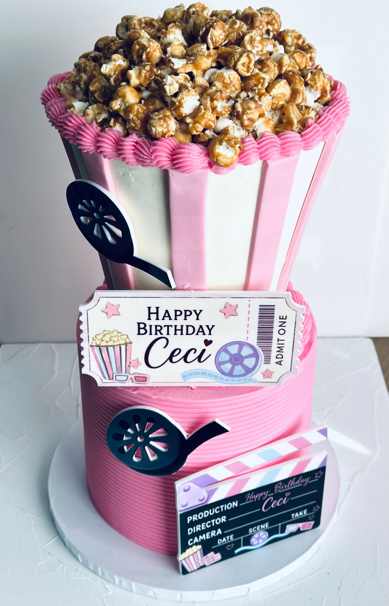Popcorn bucket with pink and white stripes, birthday message, and popcorn on top, on a white surface.