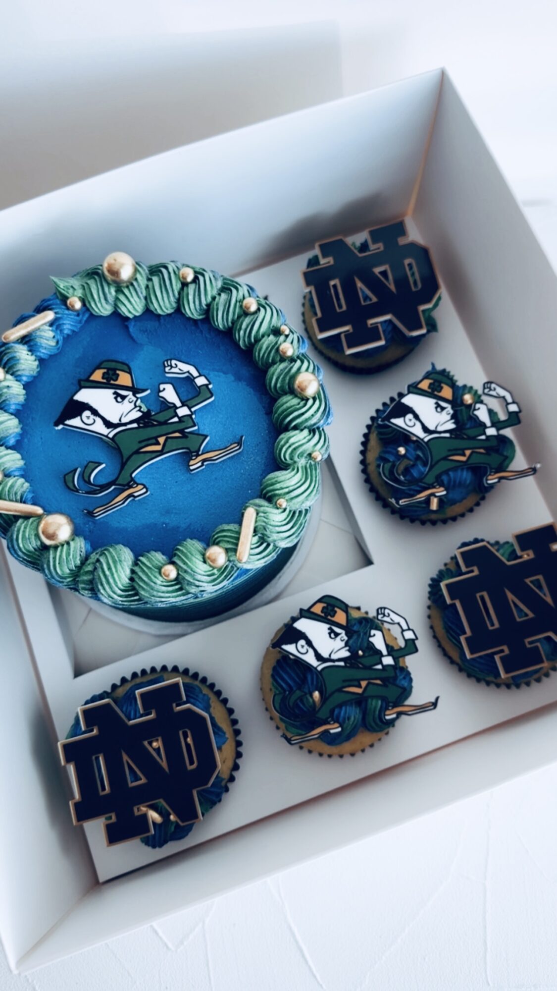 Box containing a cake and cookies with Notre Dame mascot and logo, decorated in blue and green, on a white background.