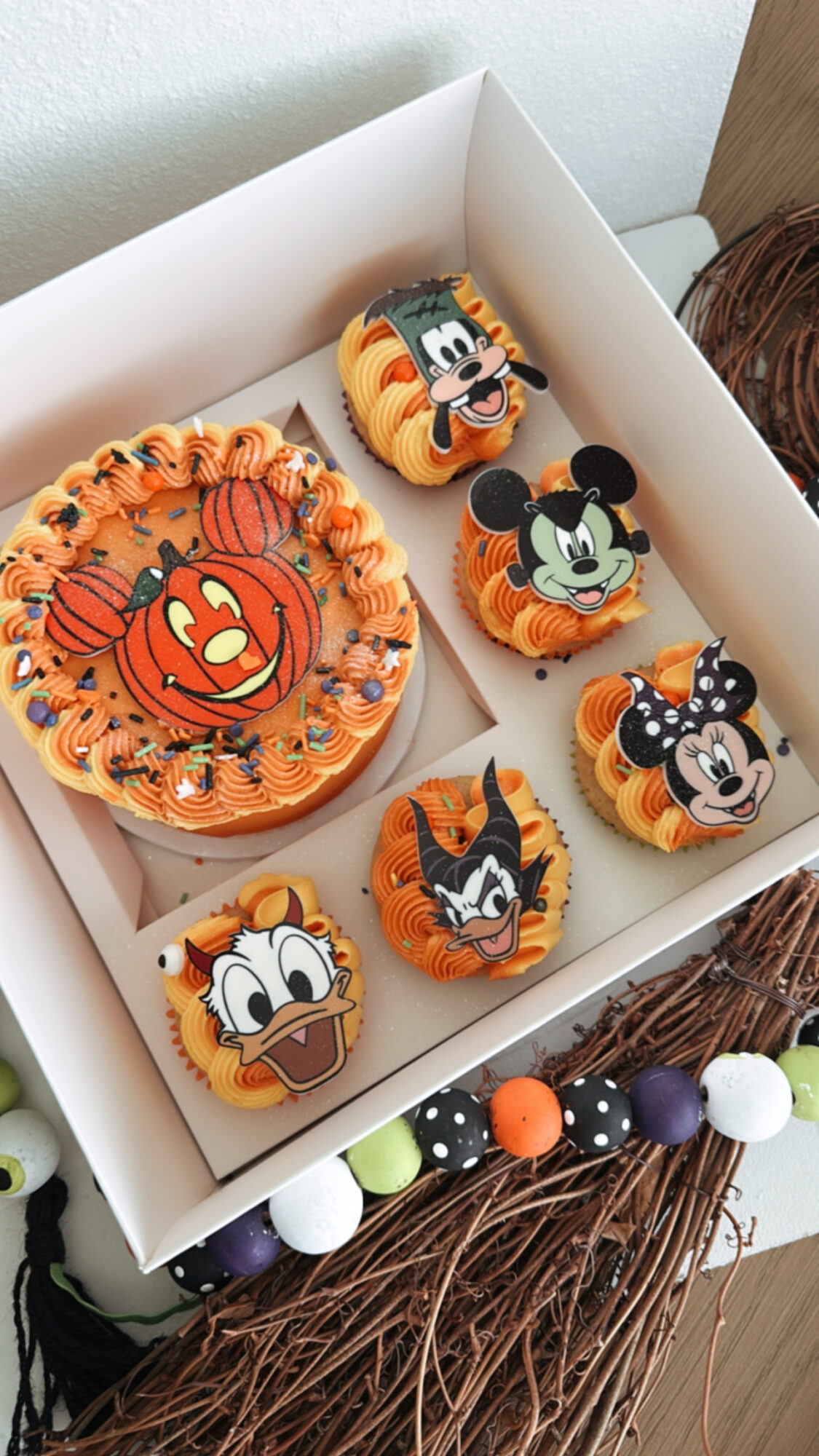 Box of six decorated cupcakes with Disney character faces, including Mickey Mouse, Minnie Mouse, and others, on a table.