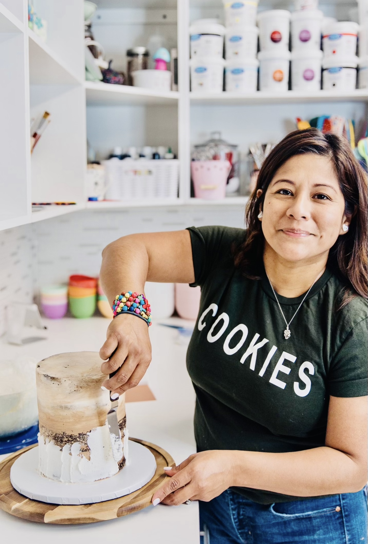 Woman with dark hair wearing a black t-shirt that says 'COOKIES' decorates a cake in a bakery or kitchen.