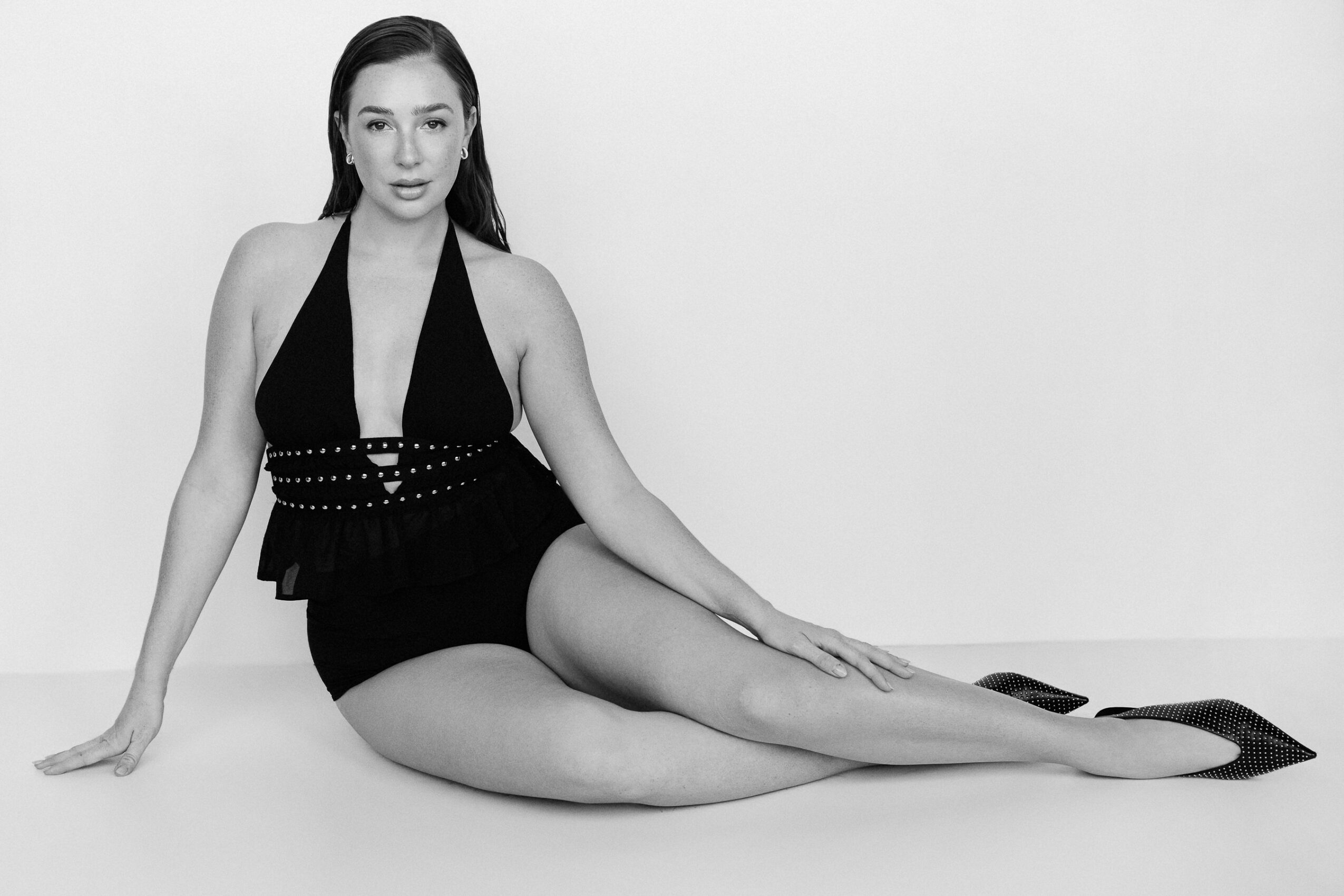 Woman sitting on the floor with legs extended, wearing a black outfit, against a plain background.