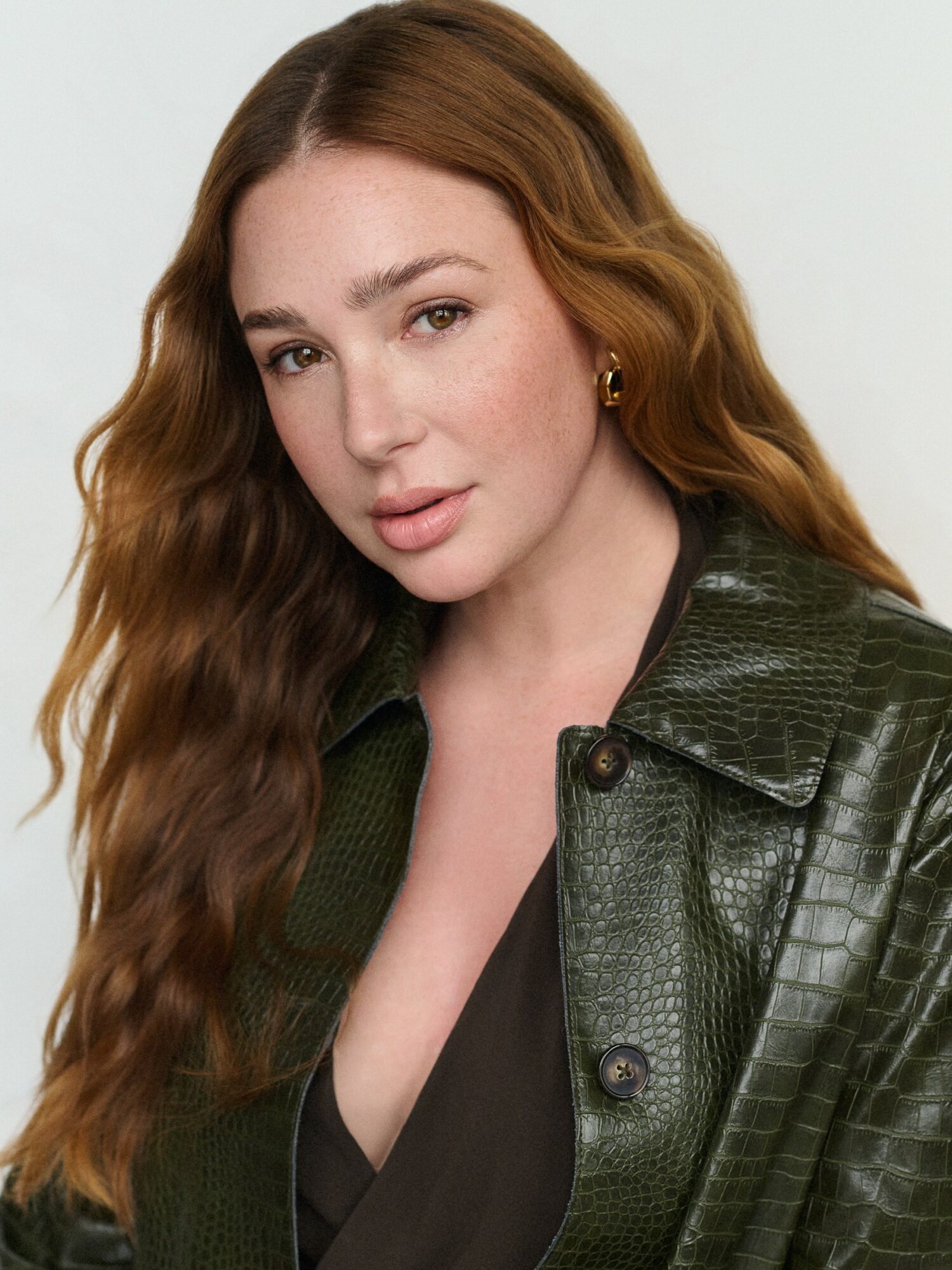 Portrait of a woman with long wavy hair wearing a dark top and green textured jacket, looking at the camera.