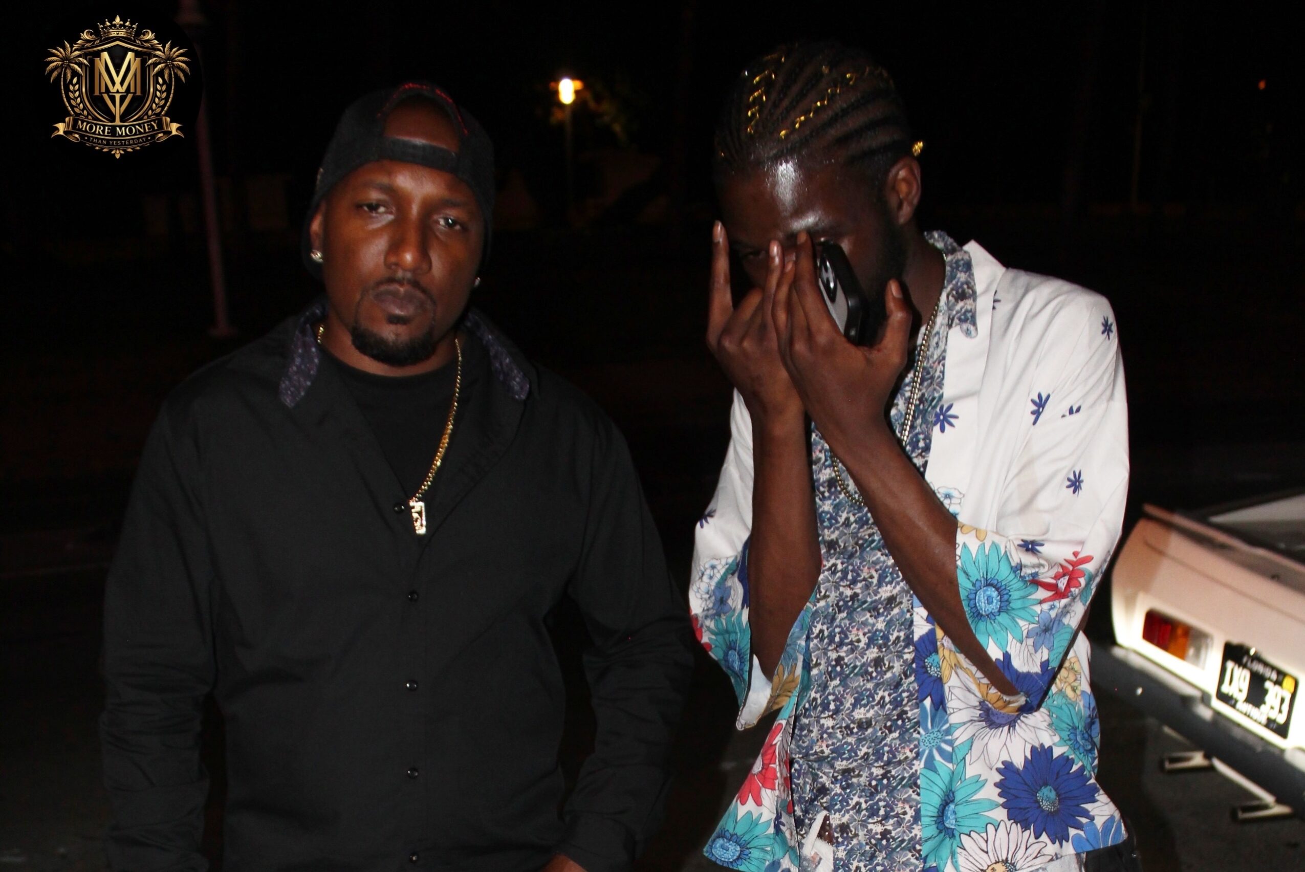 Two men standing outdoors at night, one with a serious expression, the other covering his face with his hand, wearing colorful clothing.