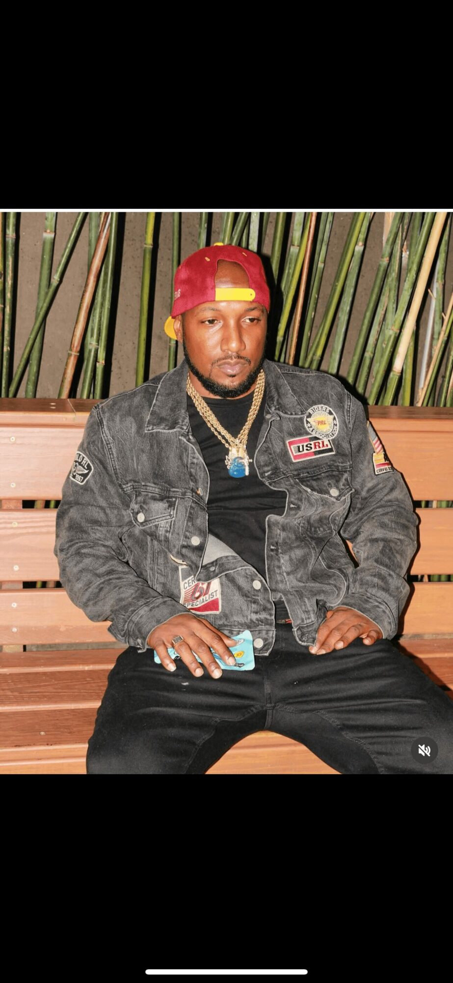 Man sitting on a wooden bench with a background of tall plants, wearing a red cap, black jacket, and gold jewelry.