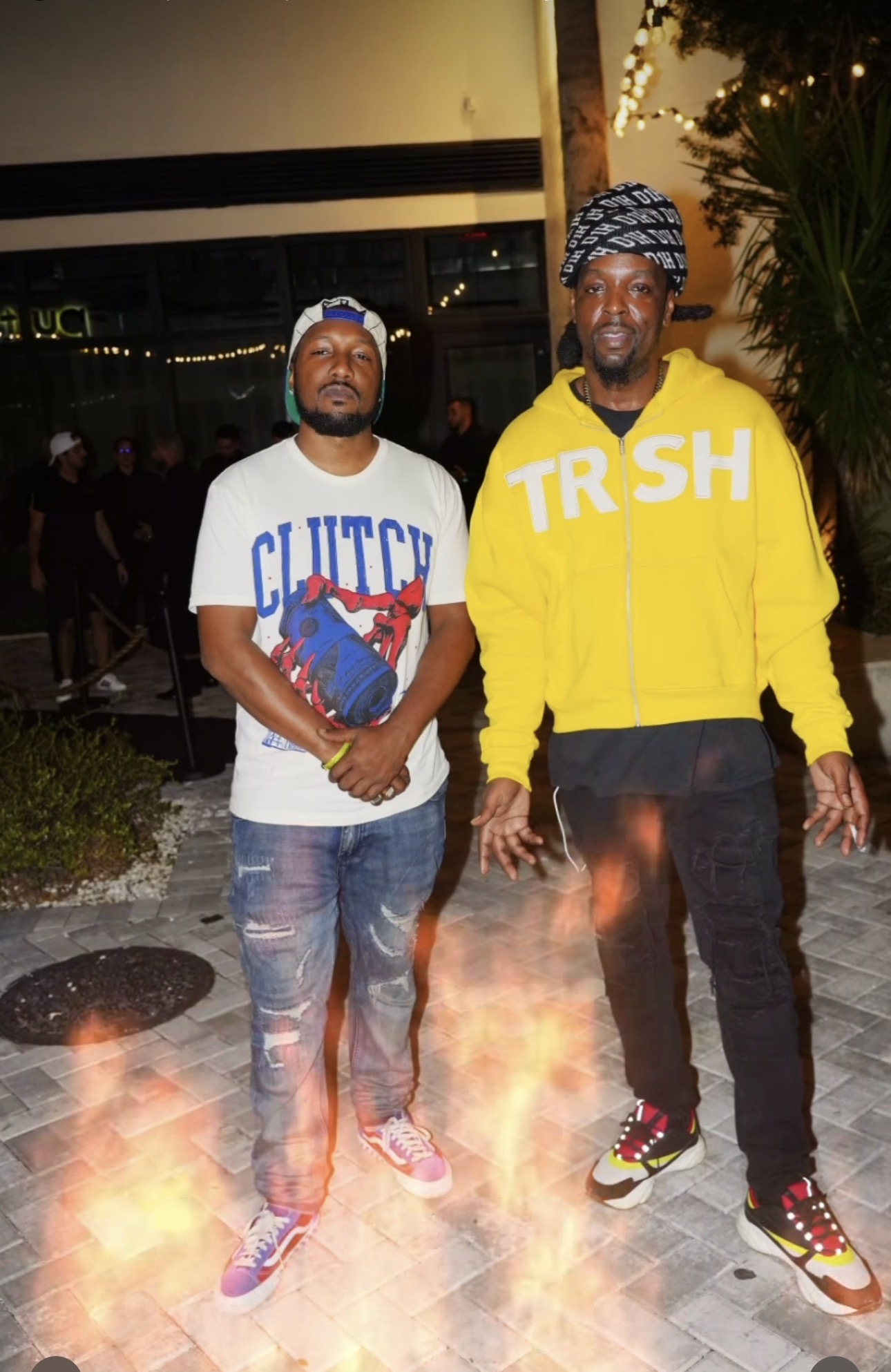 Two men standing outdoors at night, one holding a book, with fire effects at their feet, and a building in the background.