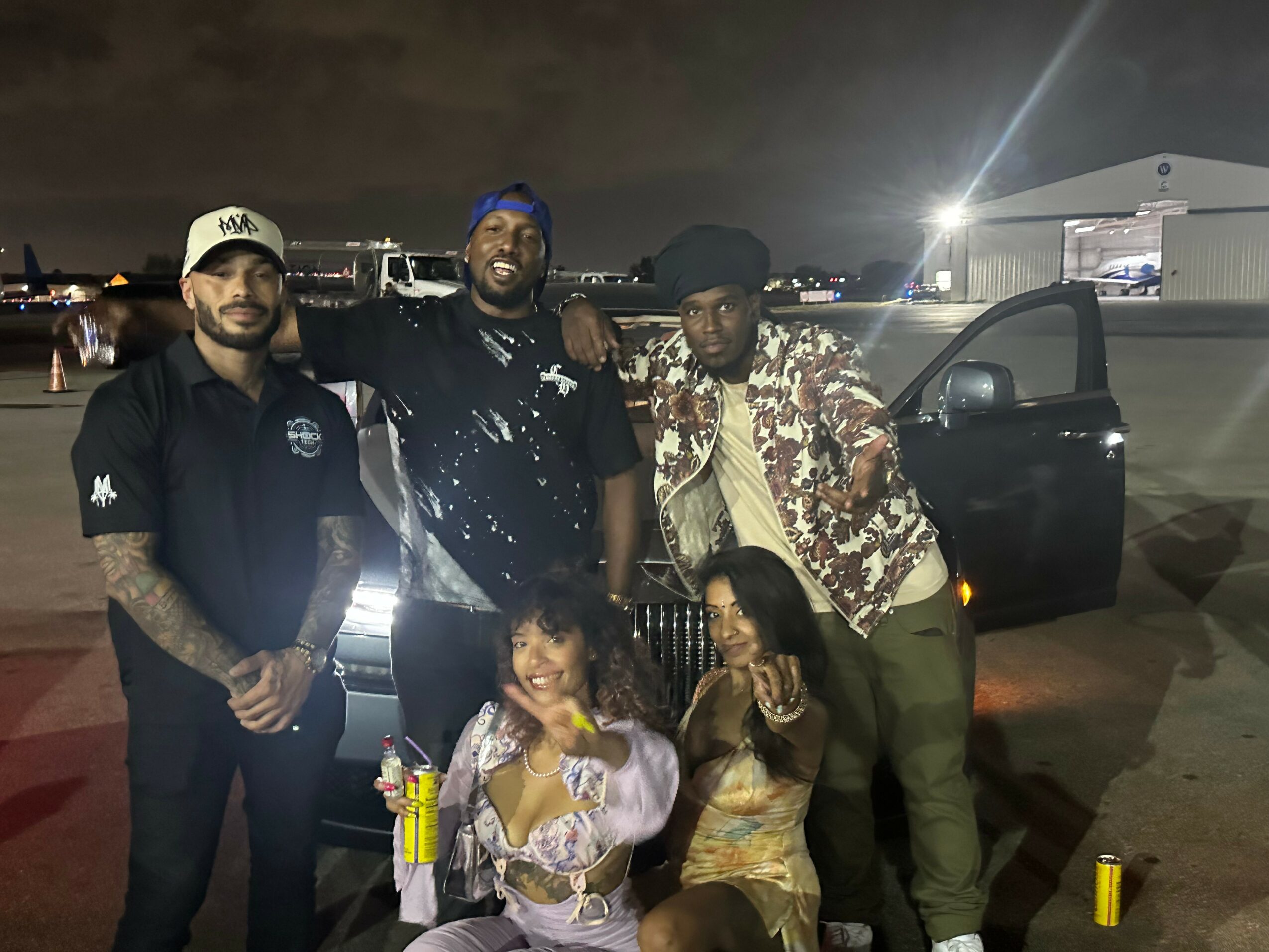 Group of five people standing outdoors at night near a car, with city lights in the background. Two women are seated in front, smiling and holding drinks. Four men stand behind them, posing for the photo.