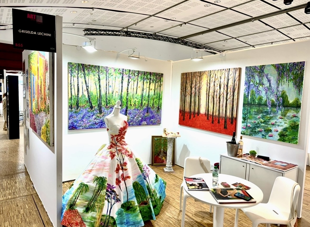 Art gallery with colorful landscape paintings on walls and a dress with rainbow-colored patterns on a mannequin, white chairs, and tables.
