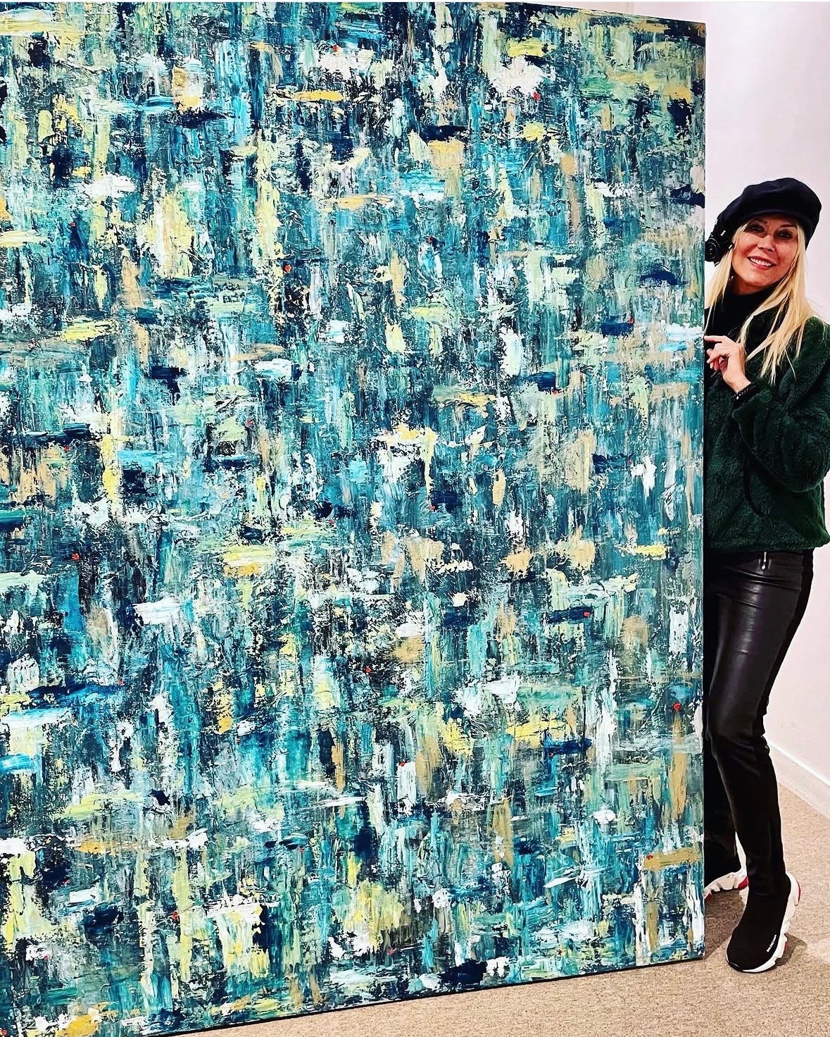 Person peeking from behind a large abstract painting with blue, green, and yellow brushstrokes.
