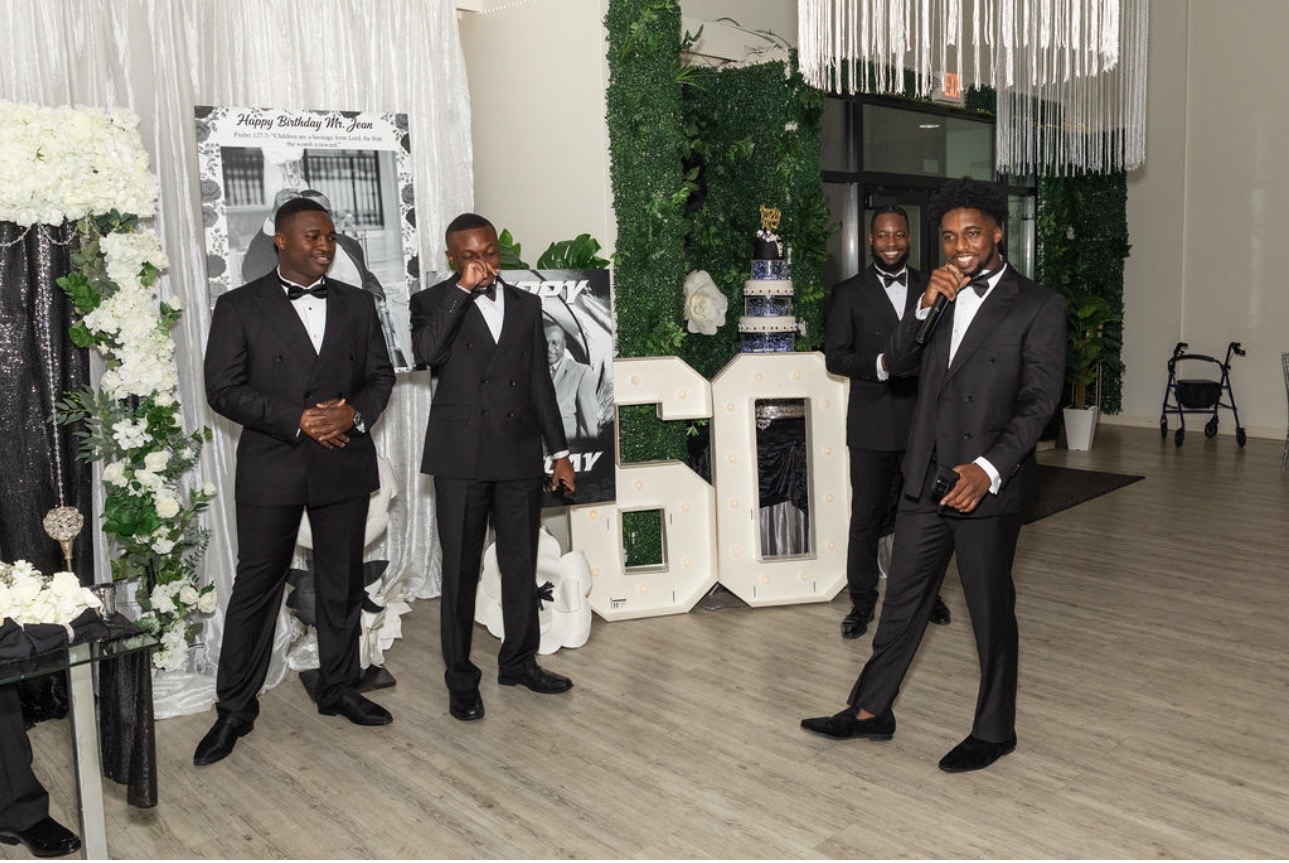Four men in tuxedos standing and talking in a decorated indoor event space with white flowers and greenery.