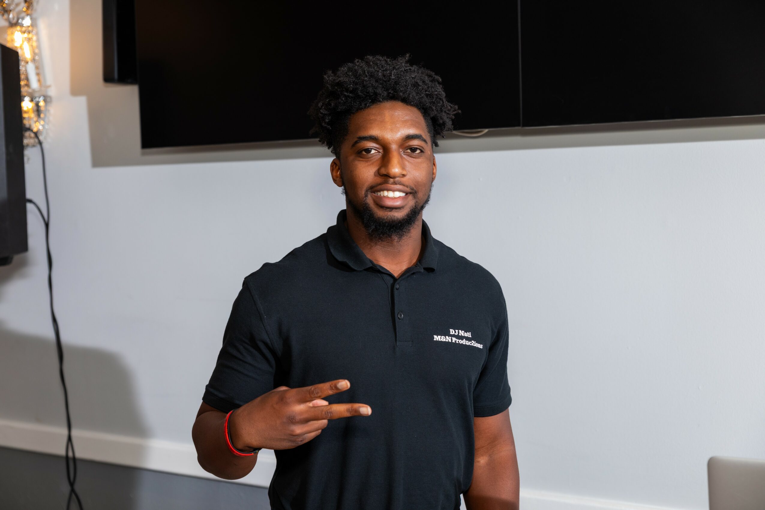 Young man with curly hair smiling and making a peace sign, wearing a black polo shirt, indoors.