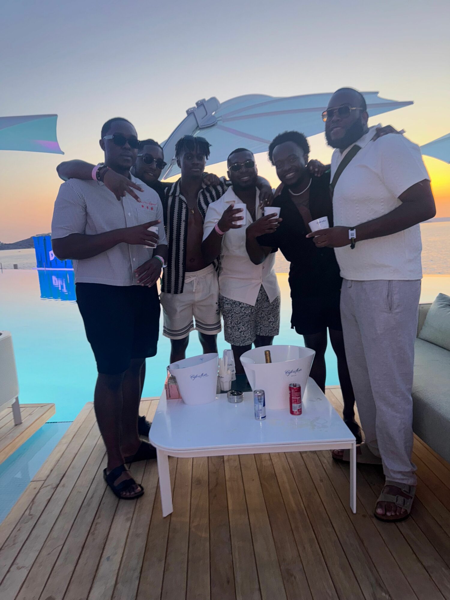 Group of six people standing around a table with drinks, on a wooden deck with ocean in background.