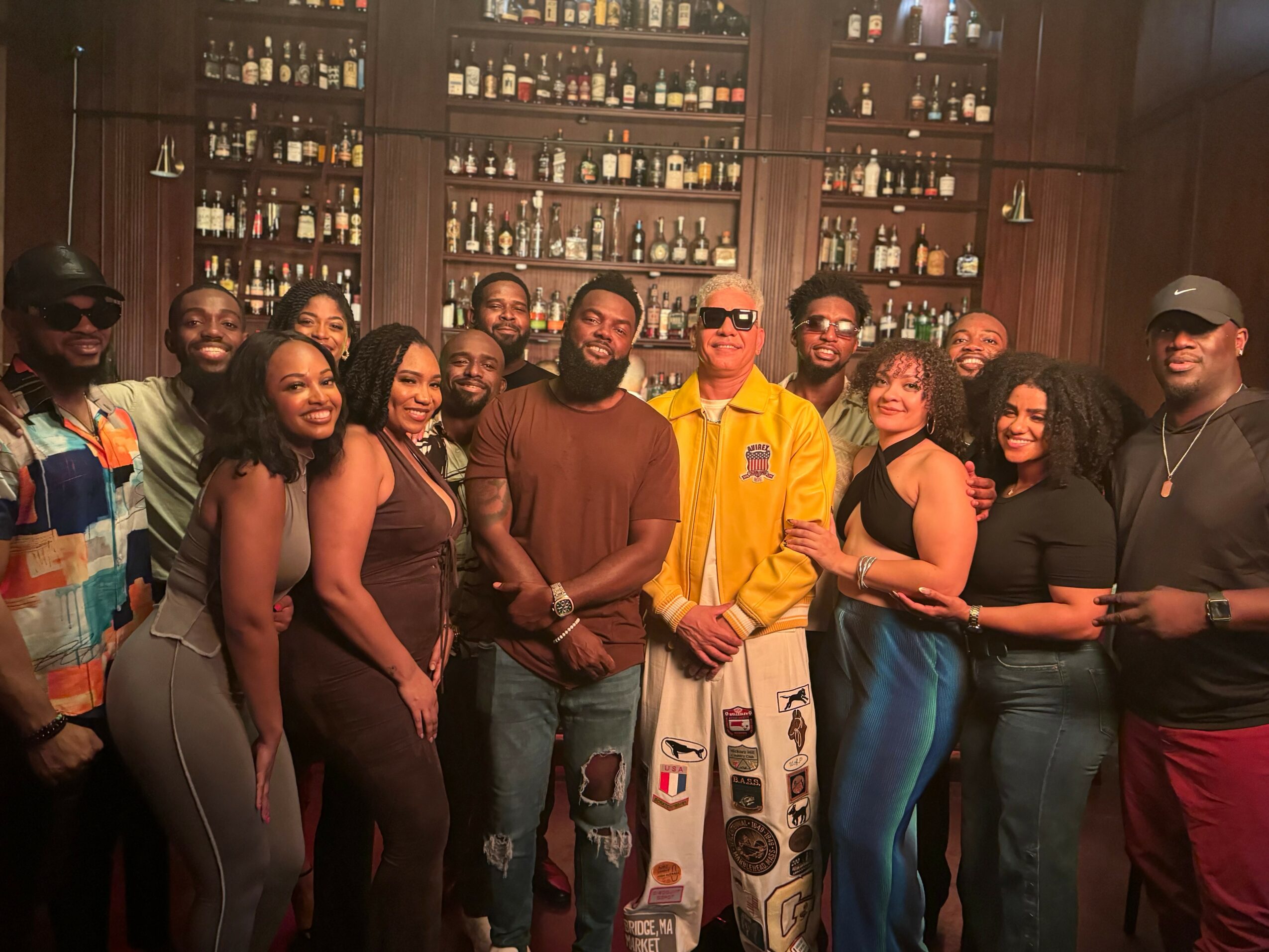Group of people posing in a bar with shelves of bottles behind them, smiling and standing close together.