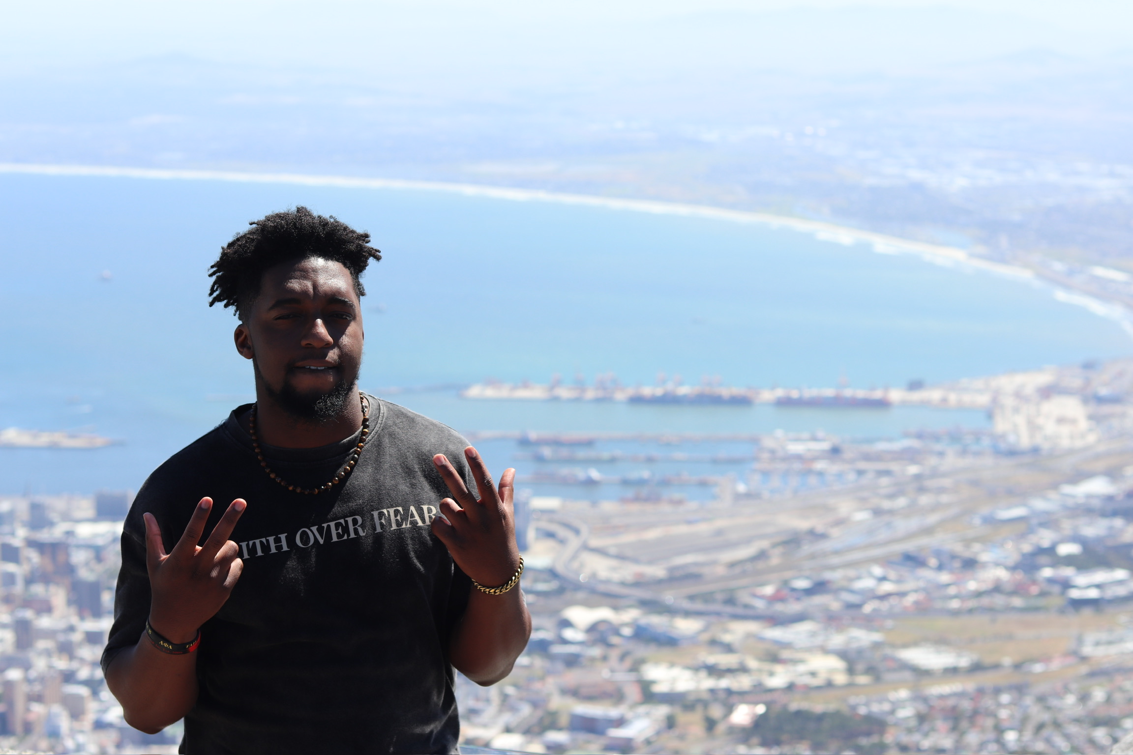 Person with dark hair and beard making hand gestures outdoors, with a coastal landscape in the background.
