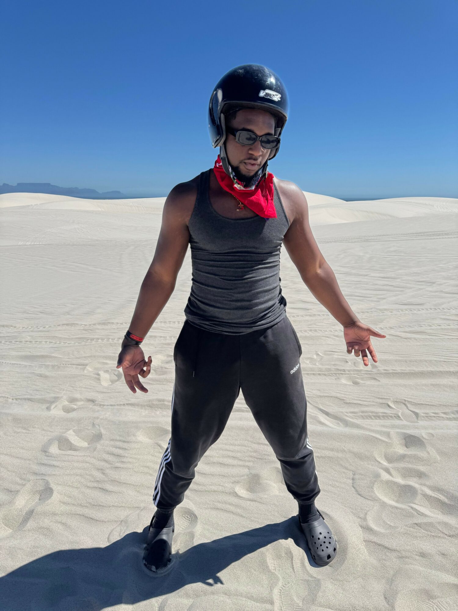Person standing on sandy beach wearing helmet, sunglasses, and casual clothing, with clear blue sky in background.